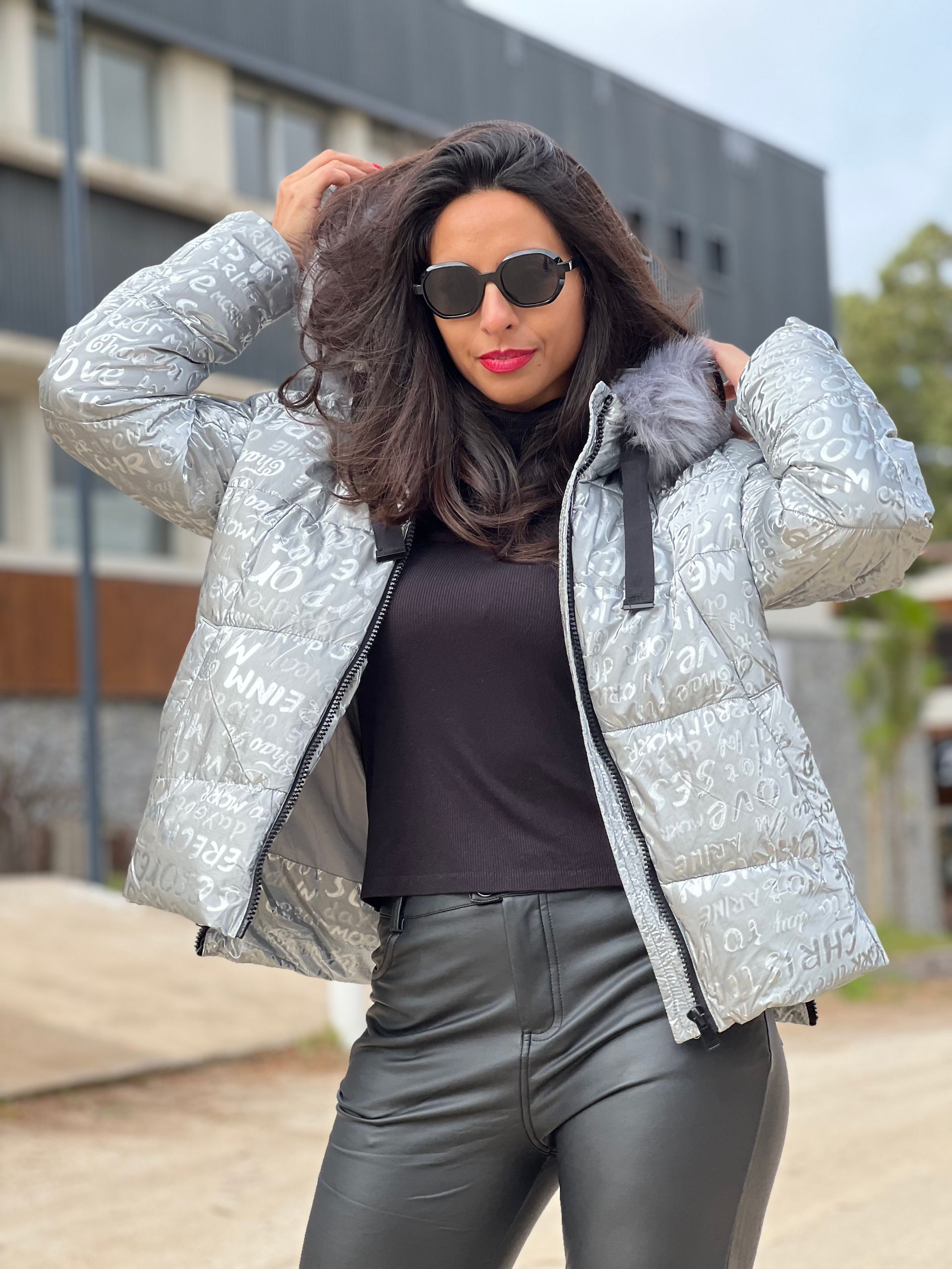 Mujer con gafas de sol ajustándose una chaqueta plateada con lentejuelas, un top negro y pantalones de cuero al aire libre.