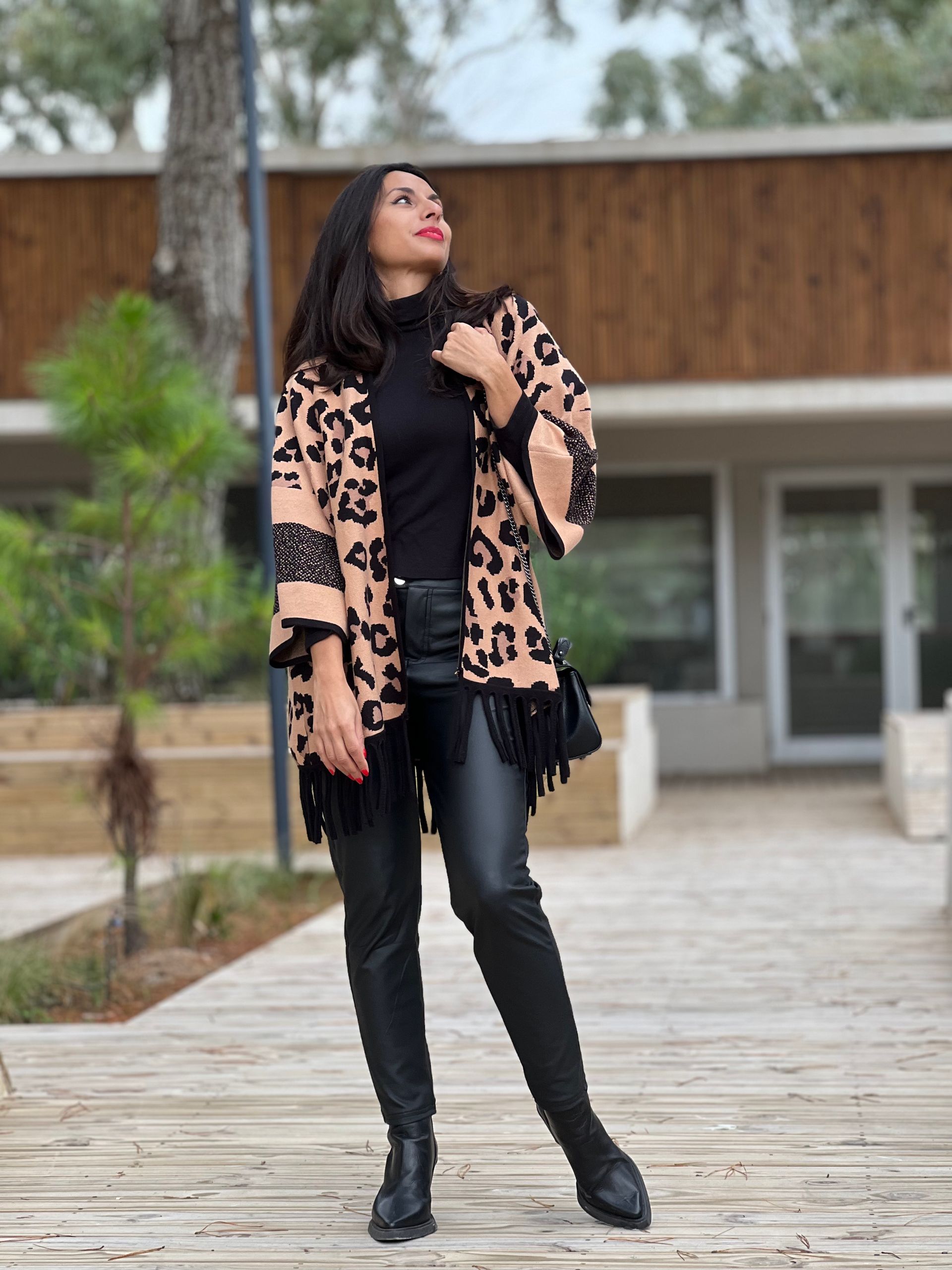 Mujer con poncho con estampado de leopardo y flecos negros, traje negro, brazos extendidos.