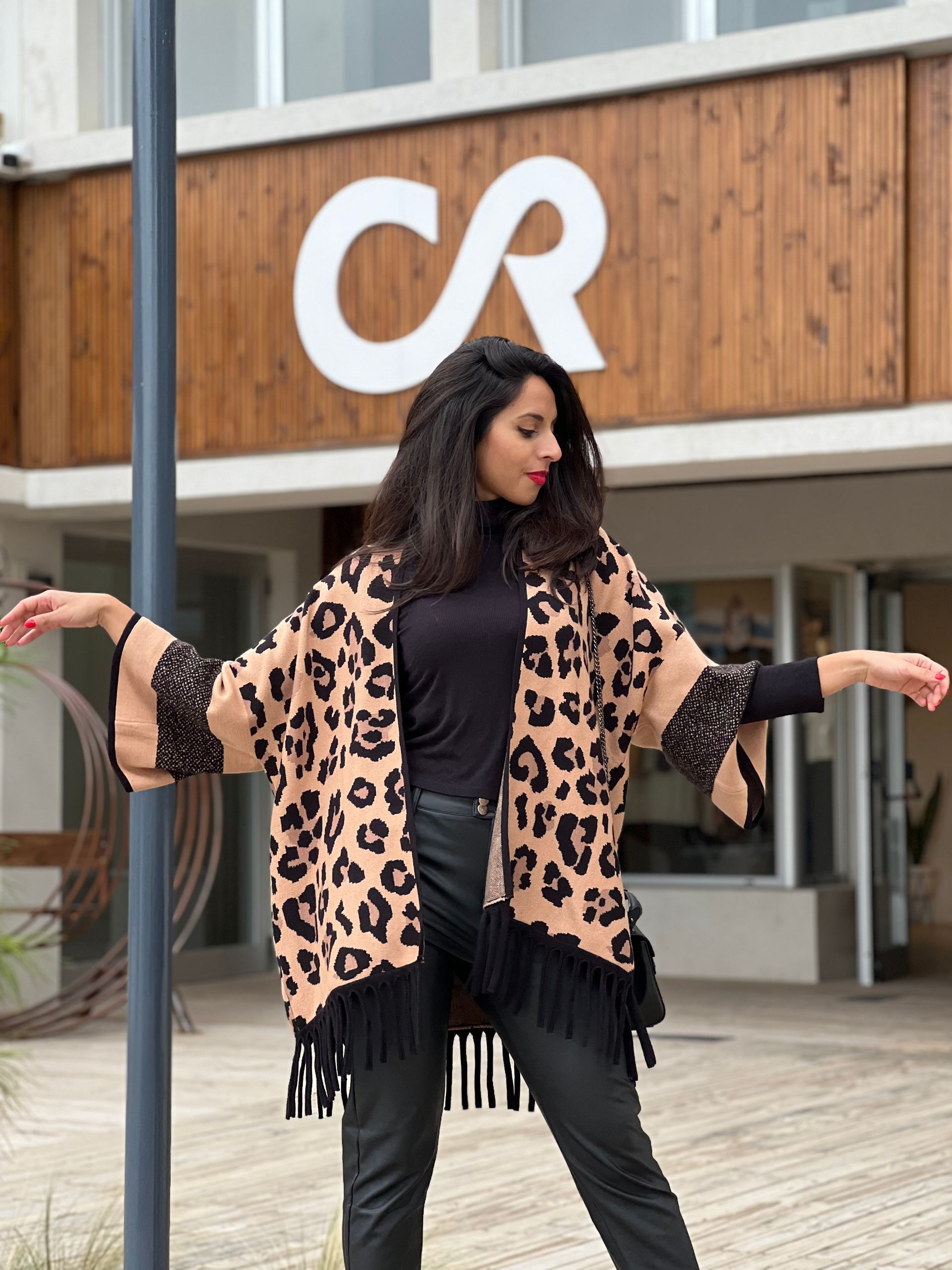 Mujer con poncho con estampado de leopardo y flecos negros, traje negro, brazos extendidos.