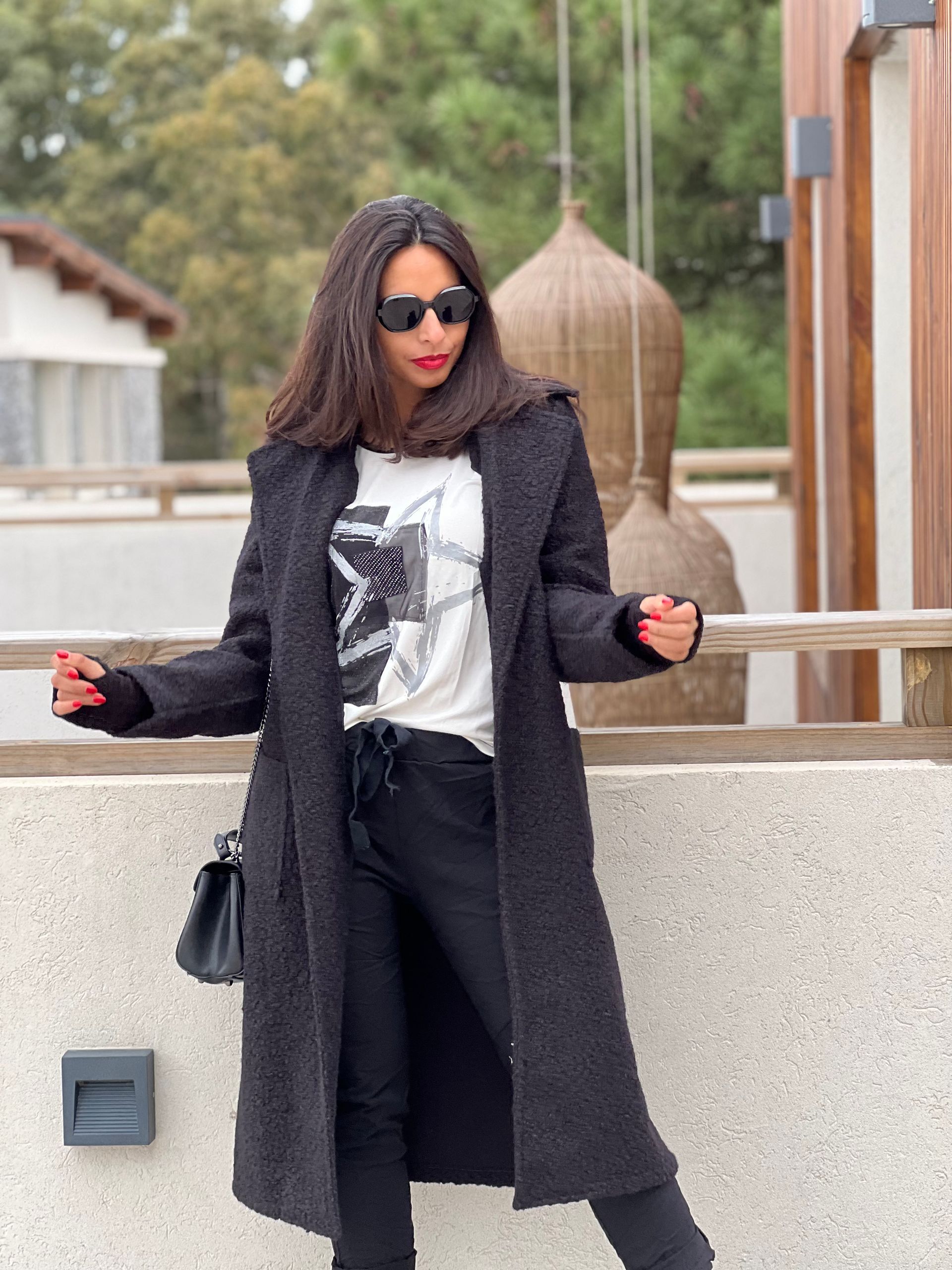 Mujer con abrigo negro y gafas de sol, de pie al aire libre, con los brazos extendidos.