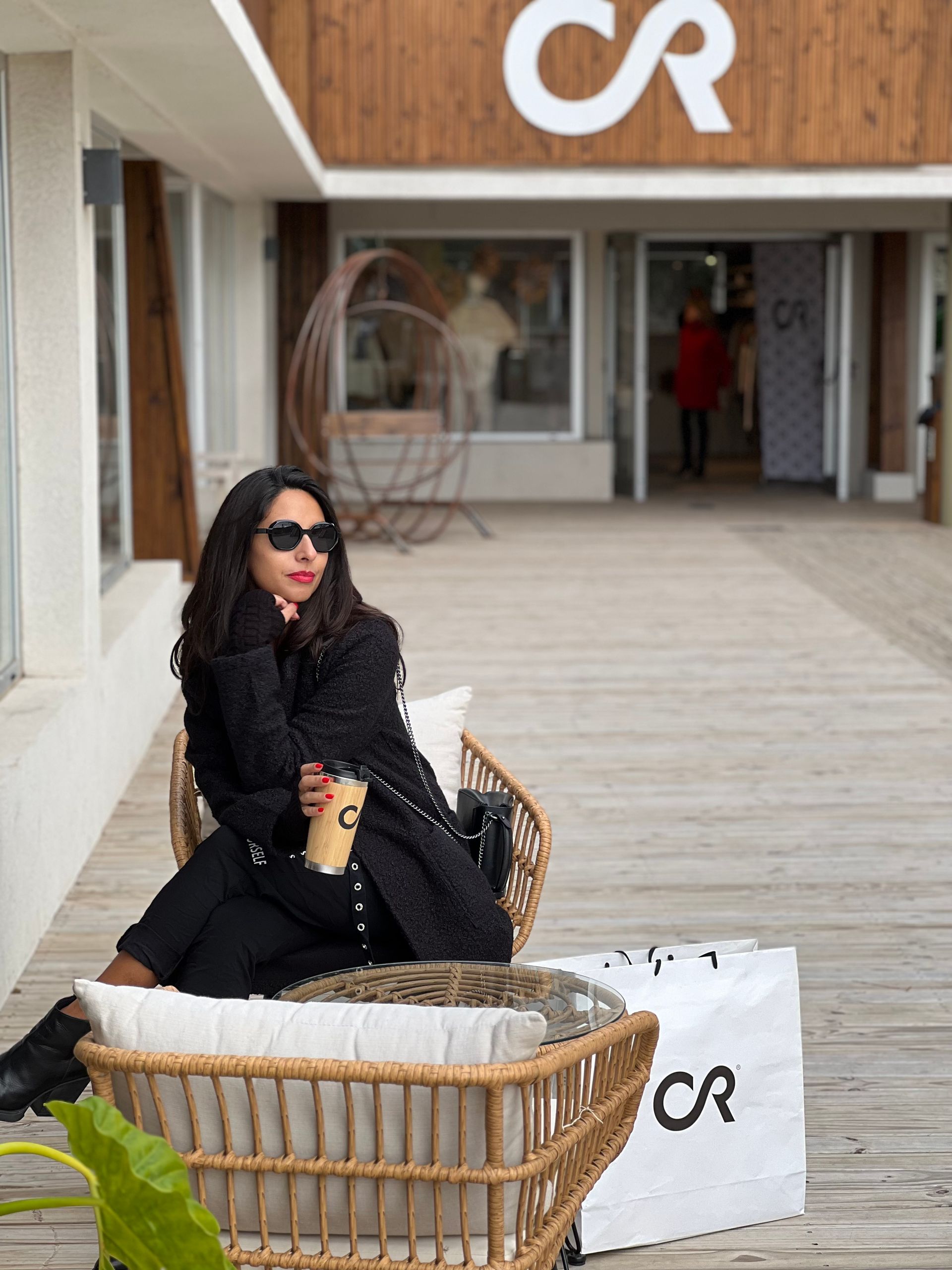 Mujer con abrigo negro y gafas de sol, de pie al aire libre, con los brazos extendidos.