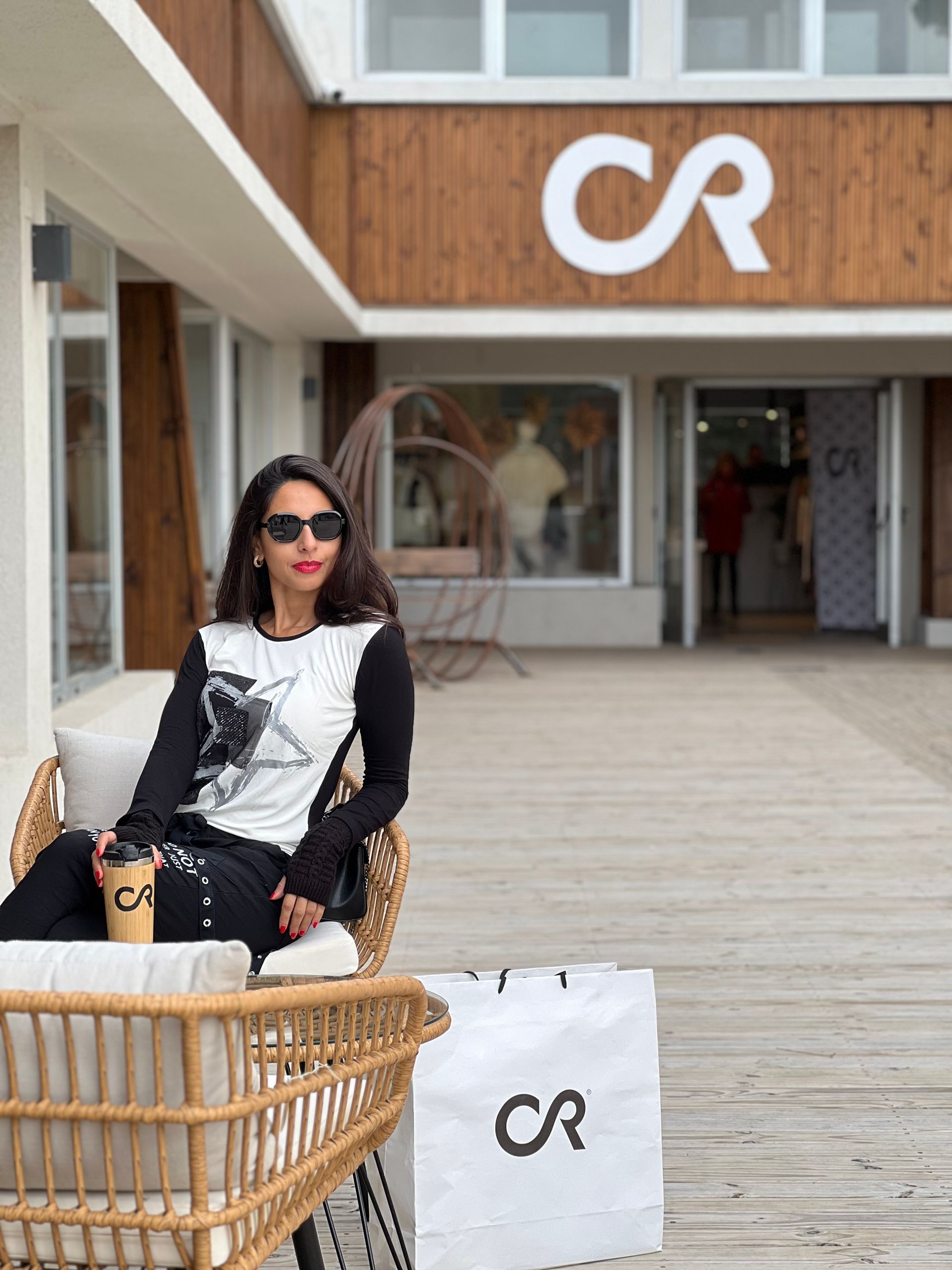 Mujer con gafas de sol sentada afuera de una tienda sosteniendo una taza de café y una bolsa de compras con el logotipo 