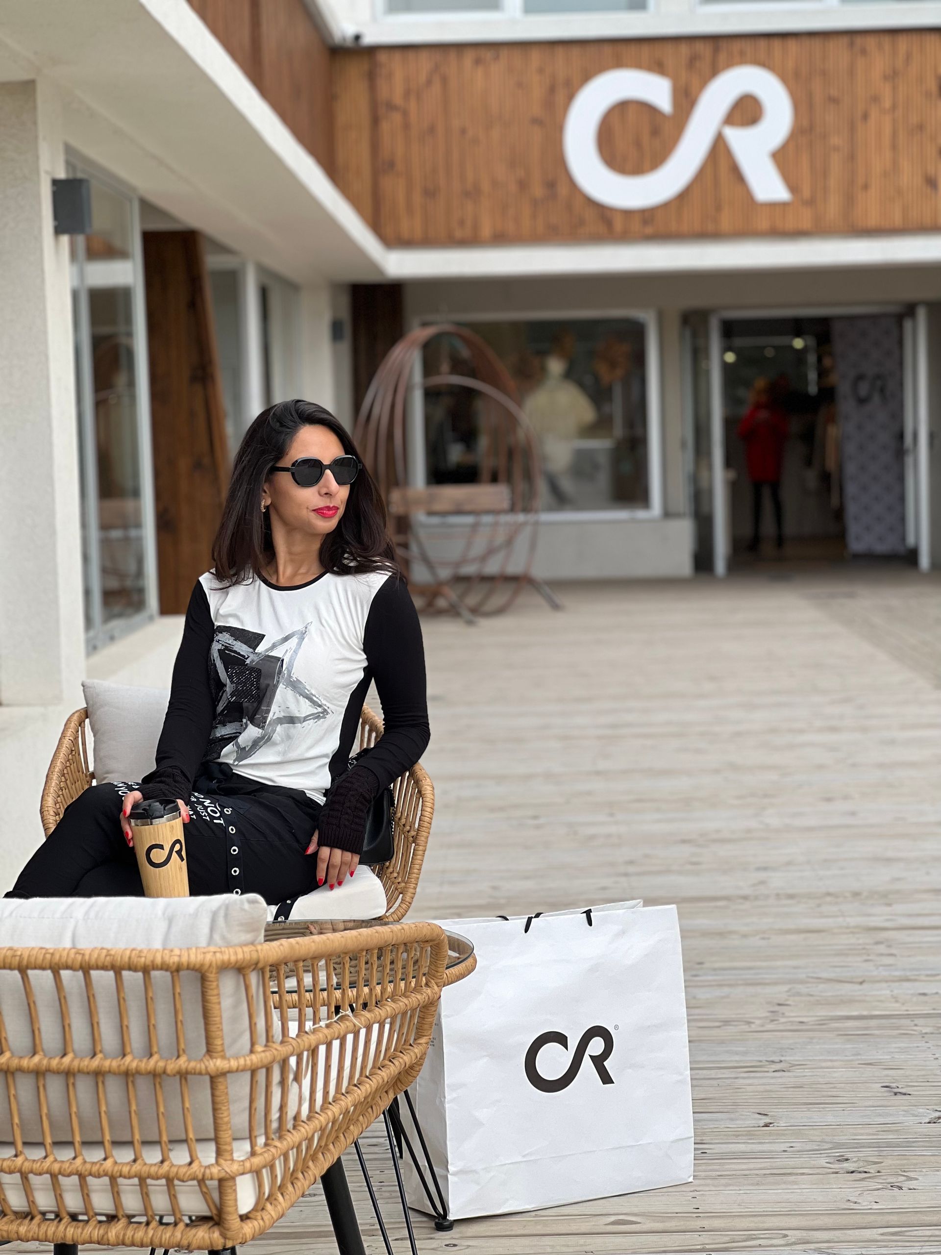 Mujer con gafas de sol sentada afuera de una tienda sosteniendo una taza de café y una bolsa de compras con el logotipo "CR".