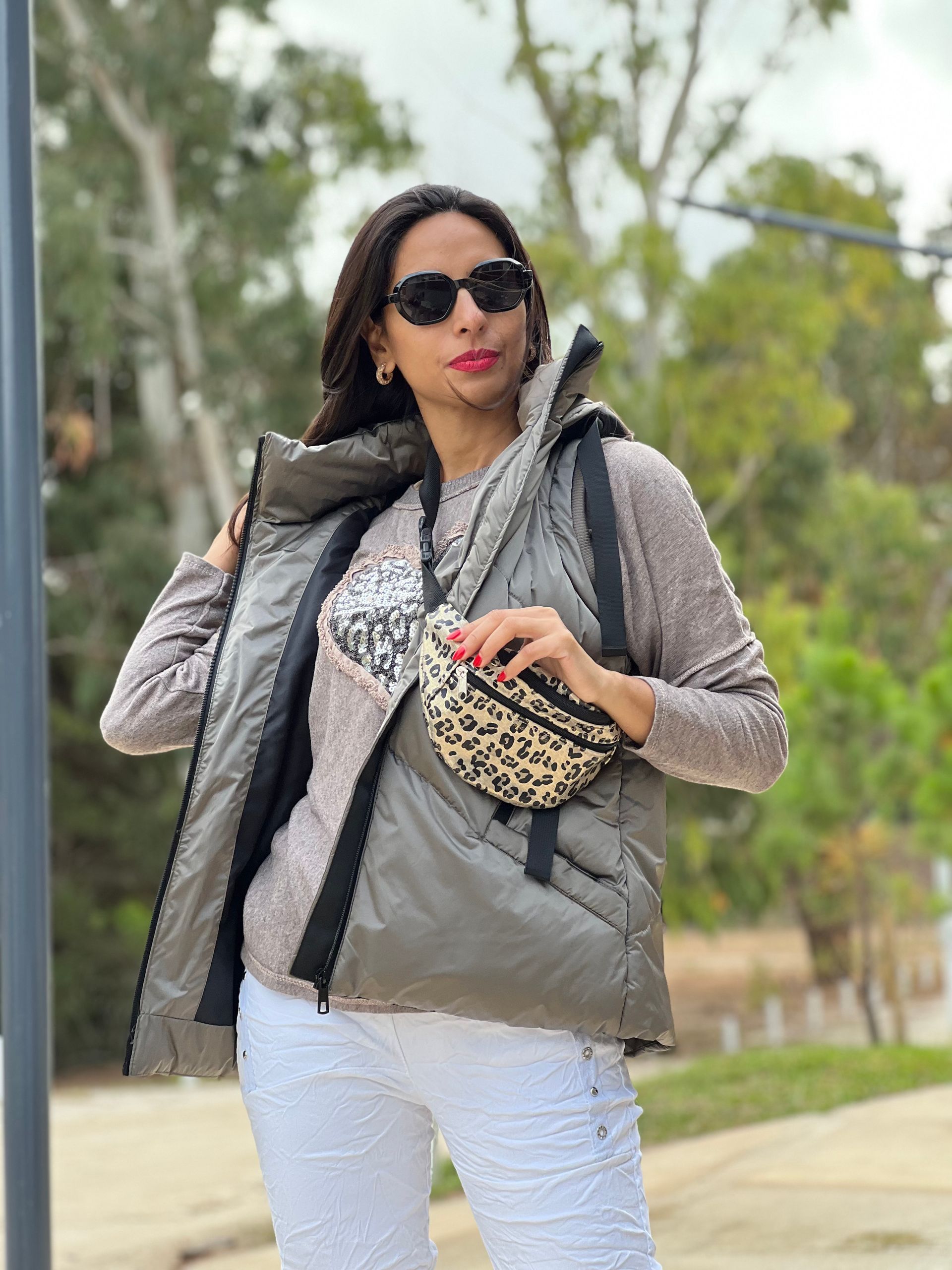 Mujer con gafas de sol, chaleco verde oliva, camisa con motivo de corazón, pantalón blanco, al aire libre.