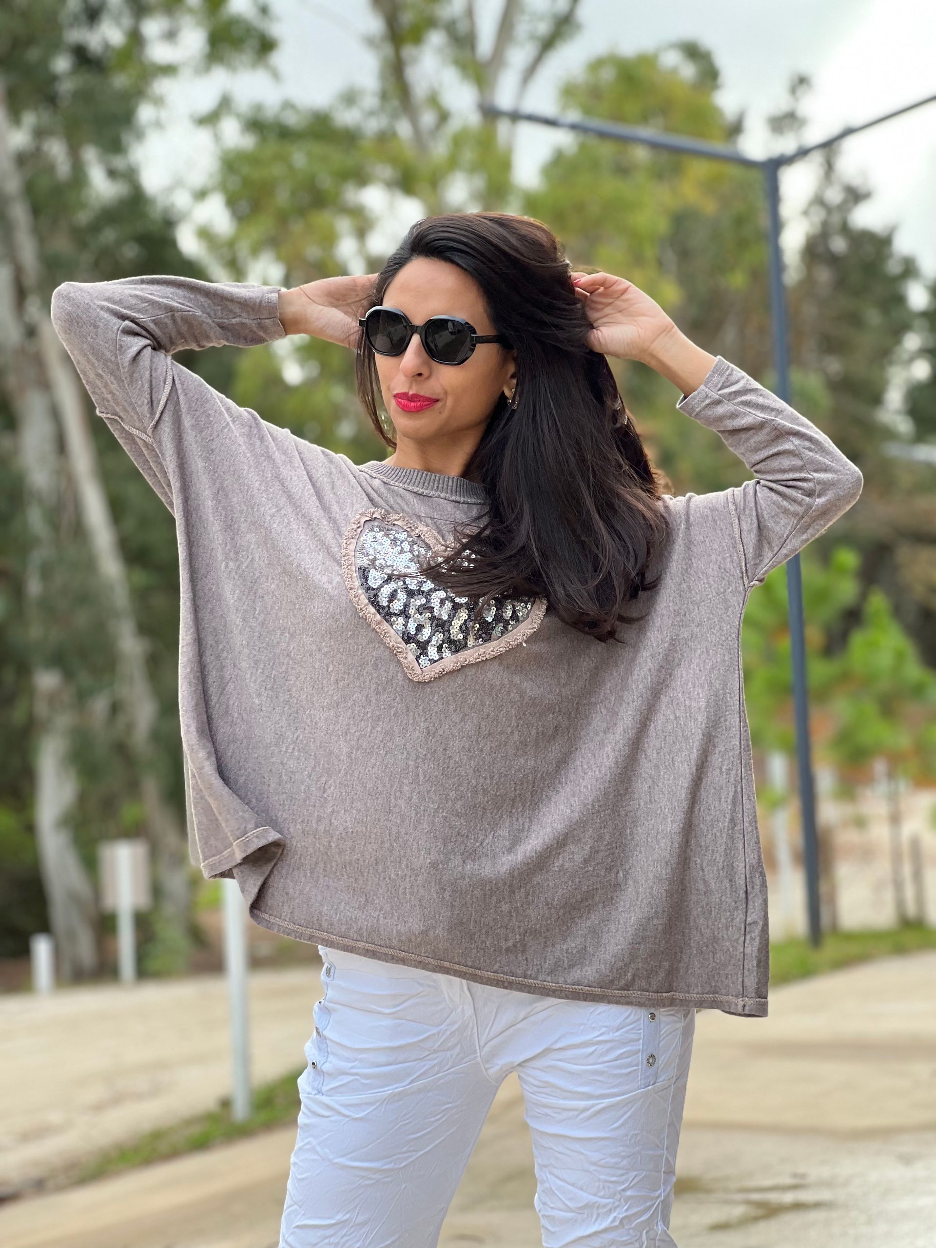 Mujer con gafas de sol, suéter gris con un corazón de lentejuelas, pantalones blancos, al aire libre.