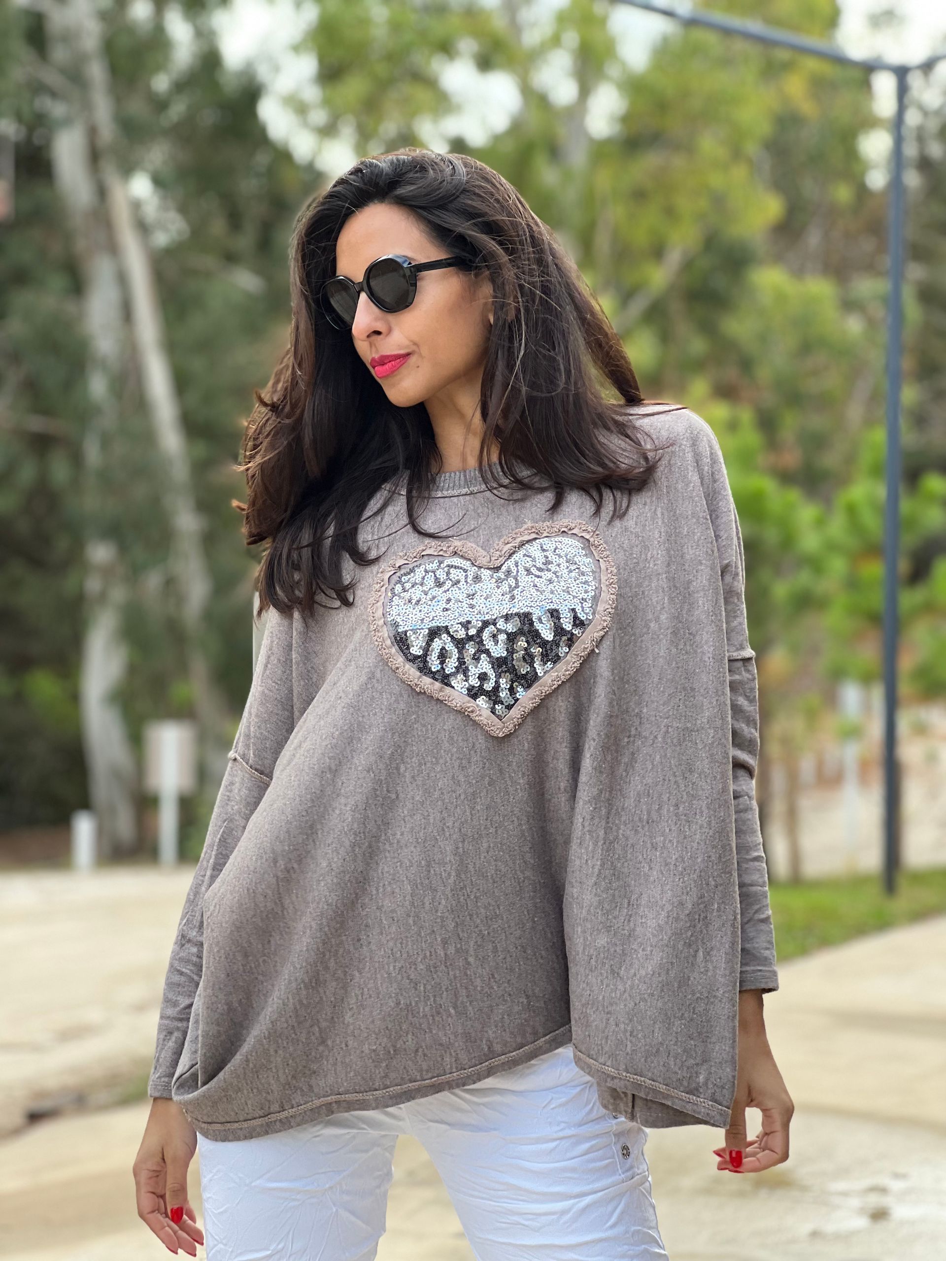 Mujer con gafas de sol, suéter gris con un corazón de lentejuelas, pantalones blancos, al aire libre.