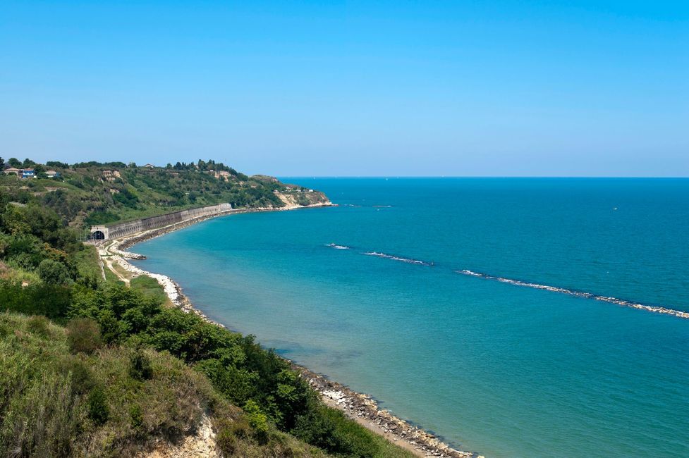 Baia costiera con acque turchesi, litorale roccioso e verdi colline sotto un cielo azzurro limpido.