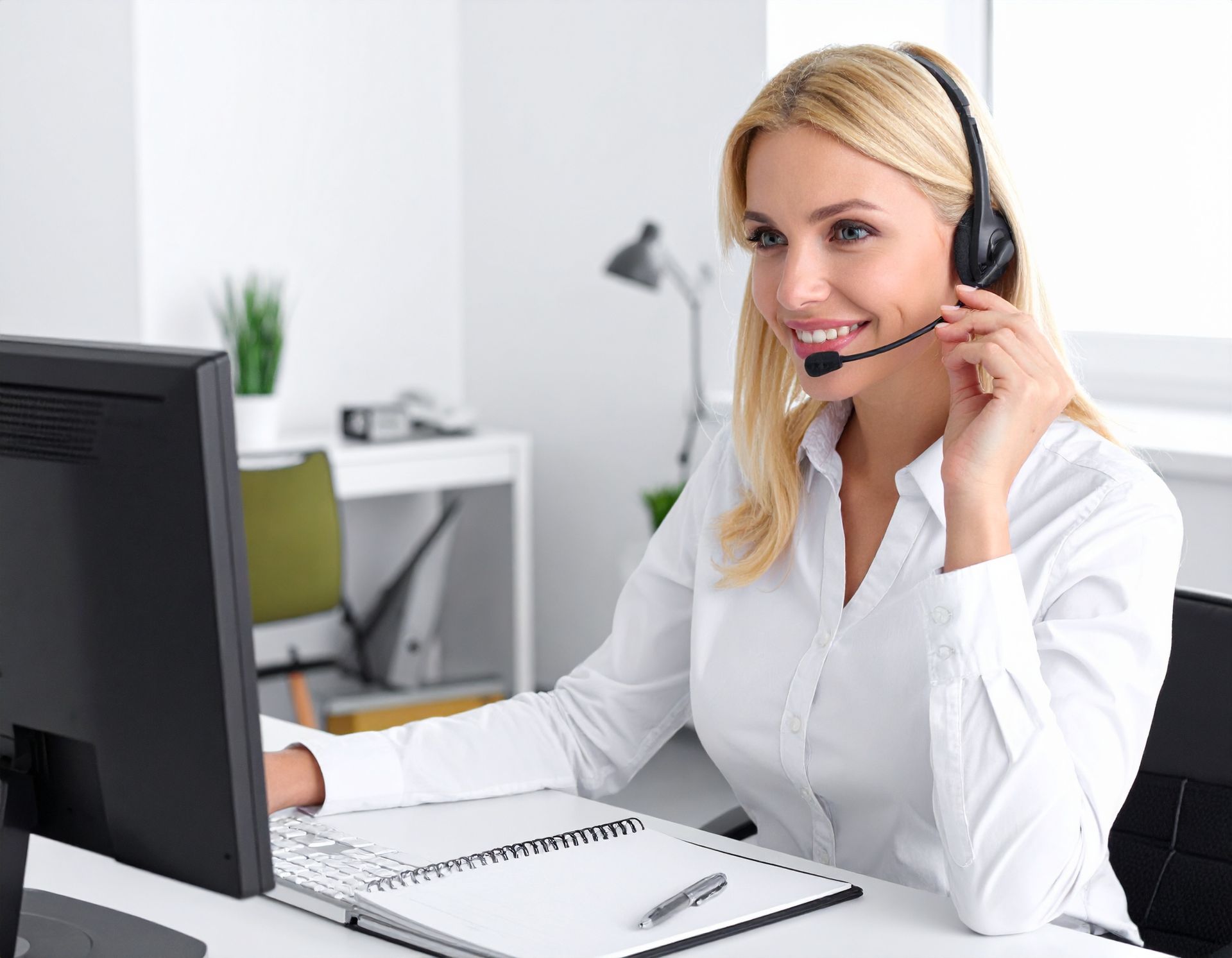 Woman with headset smiling, working at computer in an office, holding pen.