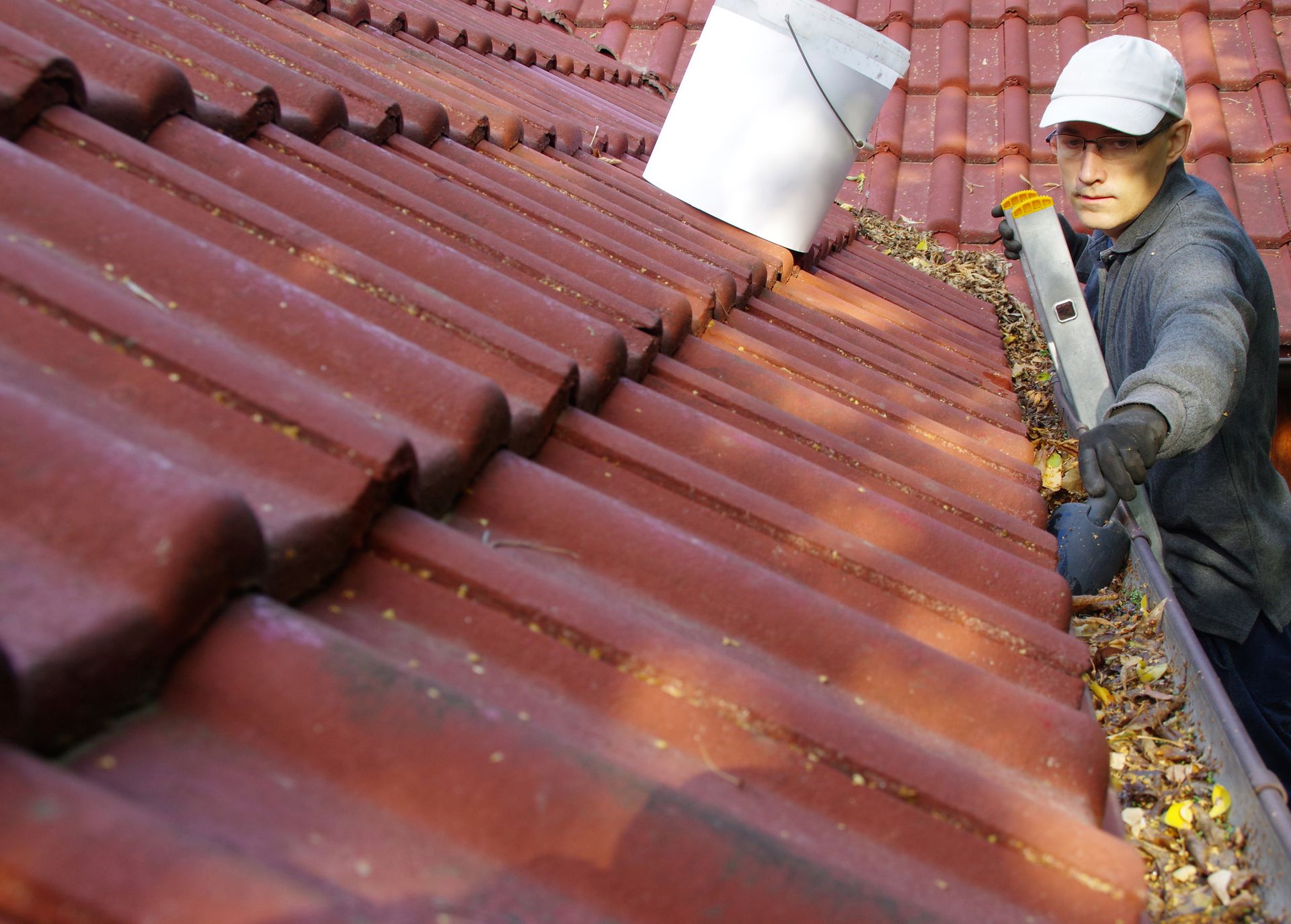 Person cleaning a gutter on a red tile roof, using a level and bucket.