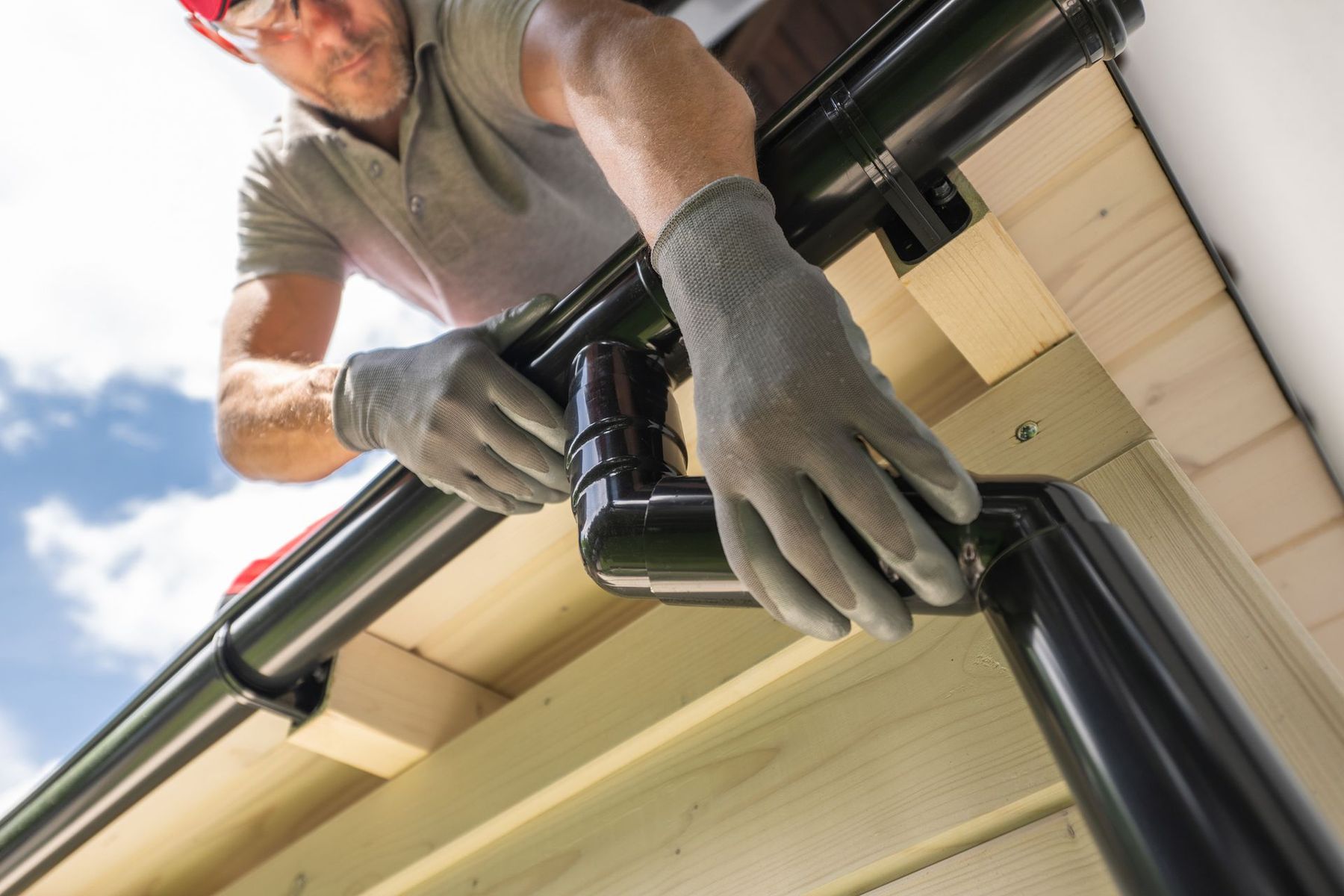 Man installing black gutter system on a building's roof with gloves, red safety glasses, and blue sky.