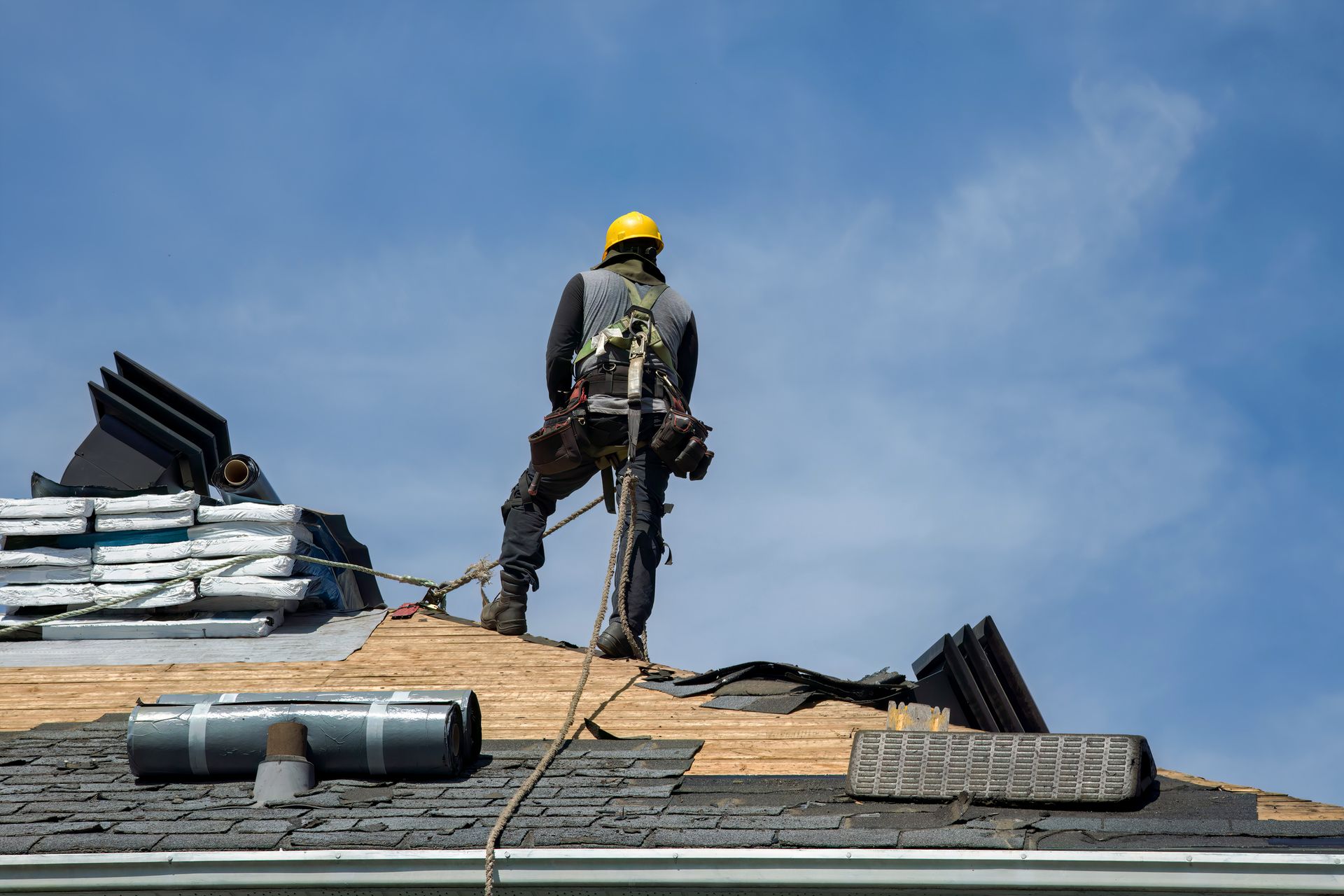 Roofer in safety harness on a roof, removing shingles under a blue sky.