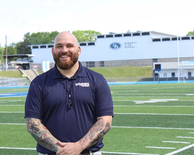 A man with a beard is wearing a suit and smiling while leaning against a wall.