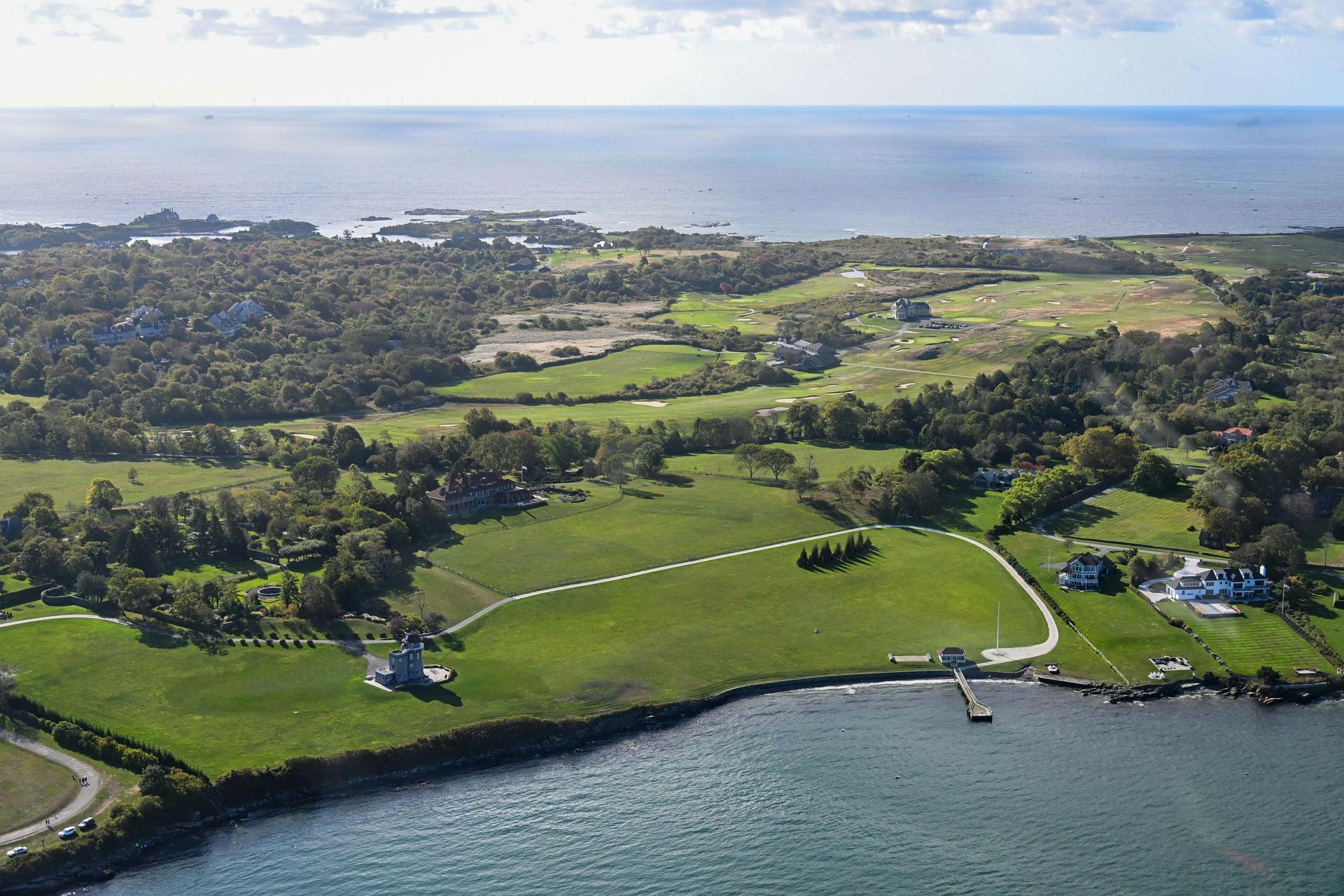 Aerial view of a coastal landscape with green fields, trees, and buildings. Ocean visible in the background.