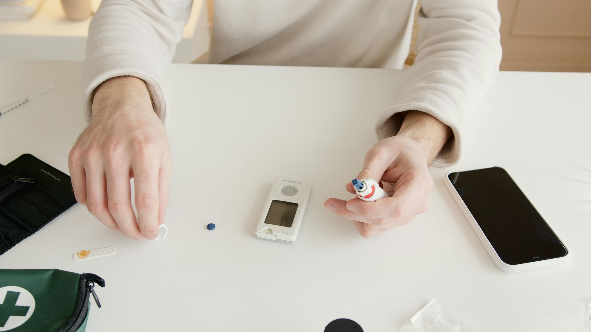 Person using blood glucose meter on white table, with supplies like a phone and first aid kit.