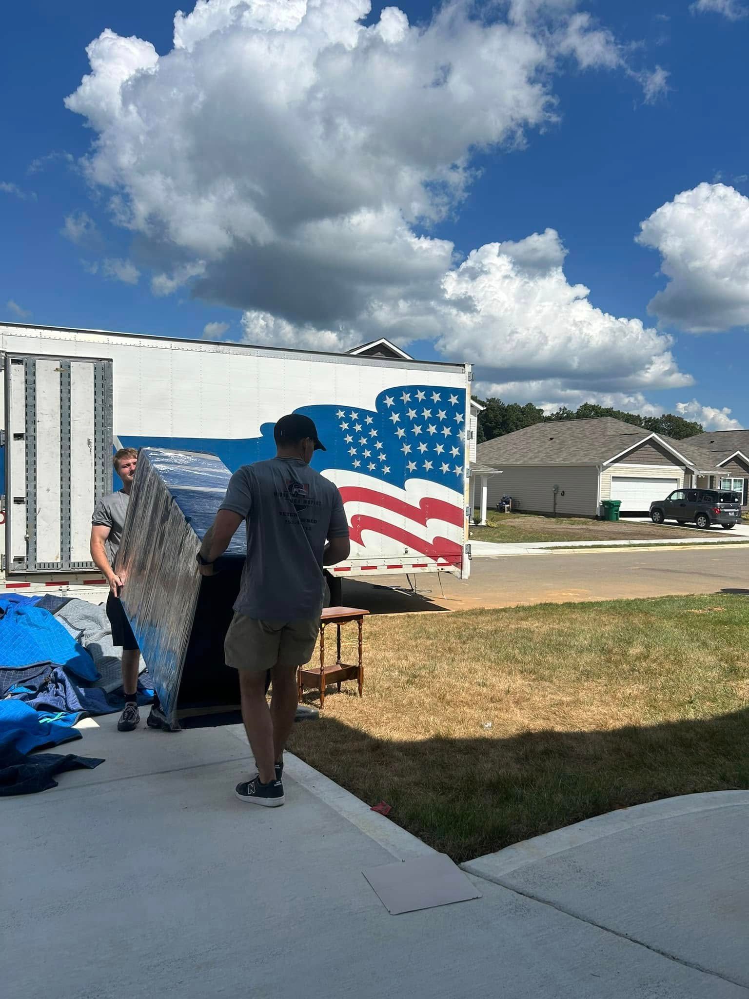 Two men loading furniture into a moving truck with an American flag on it under a blue sky.