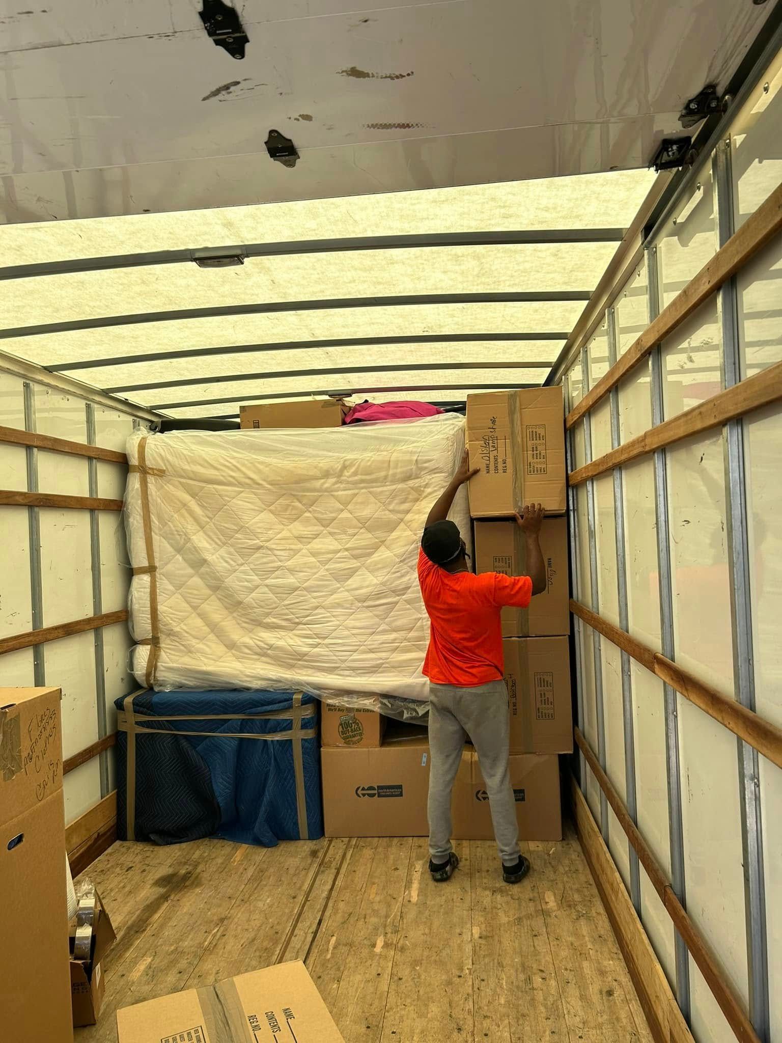 Man in orange shirt loading boxes inside a truck, other items and a mattress visible.