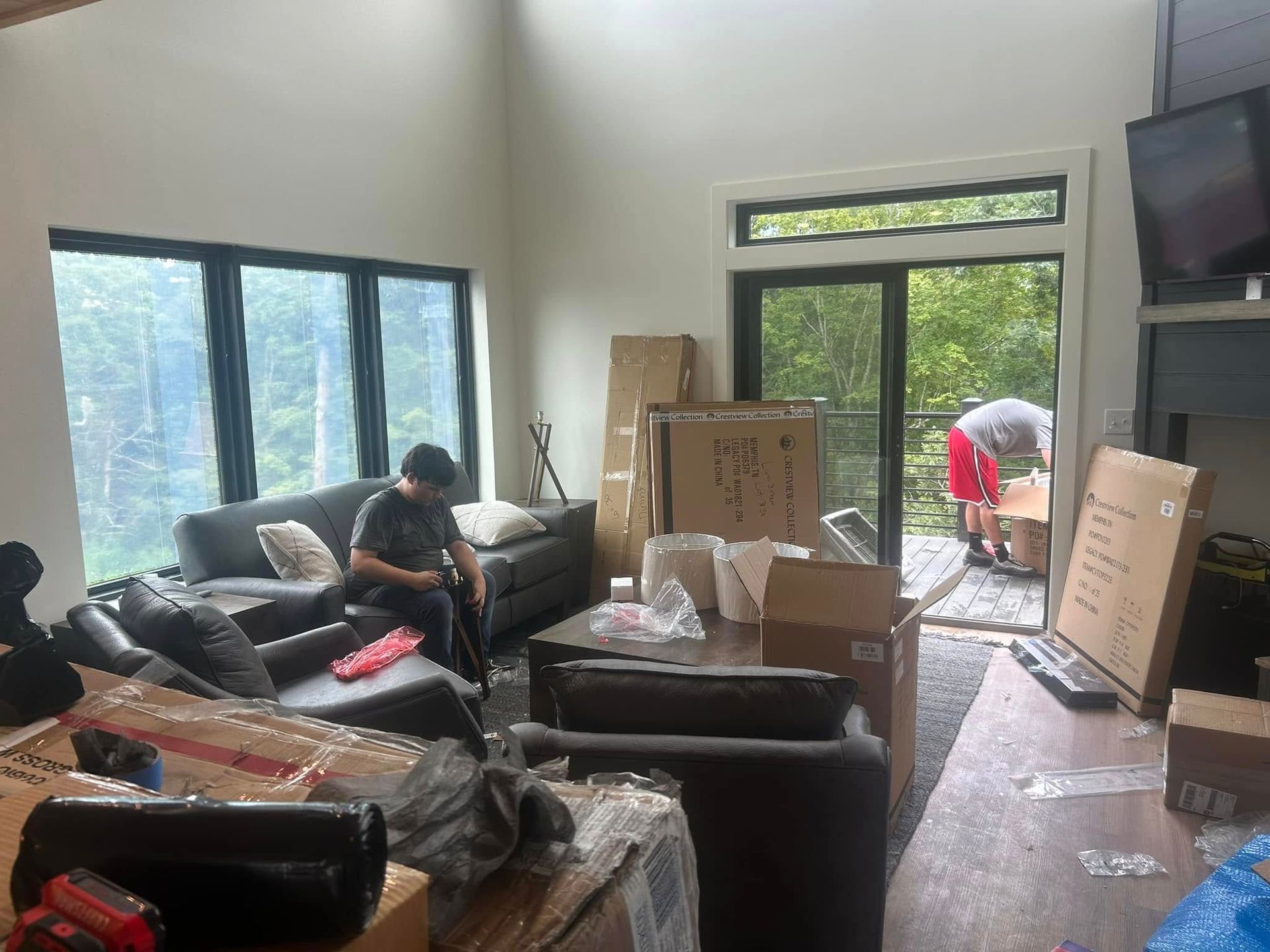 Living room with furniture, boxes, and a person on the couch. Another person outside by the sliding door with boxes.
