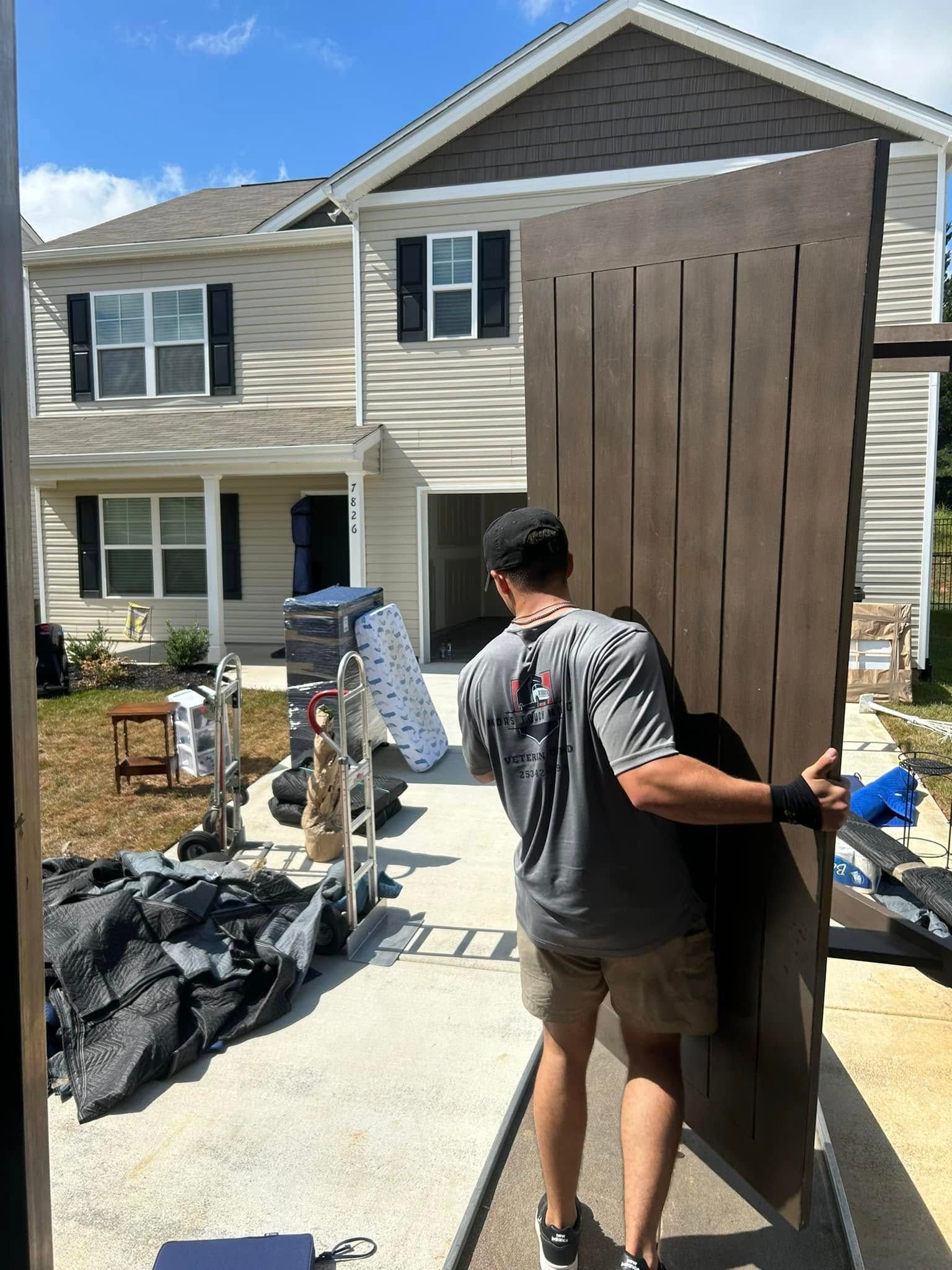 Man carrying a large brown wooden door towards a house, during a move. House and moving supplies in the background.