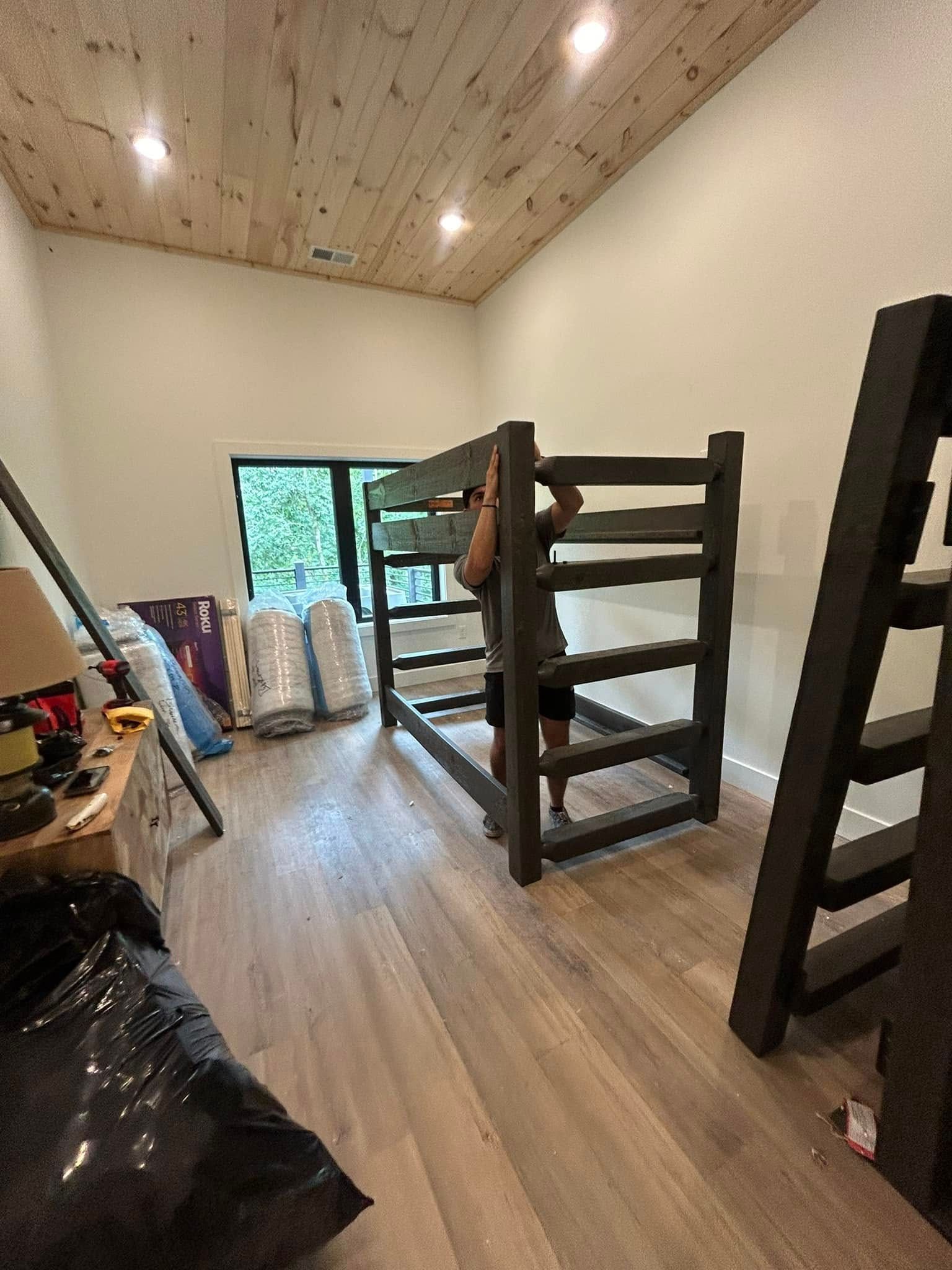 Person assembling a dark-stained bunk bed in a room with light-colored walls and wood flooring.