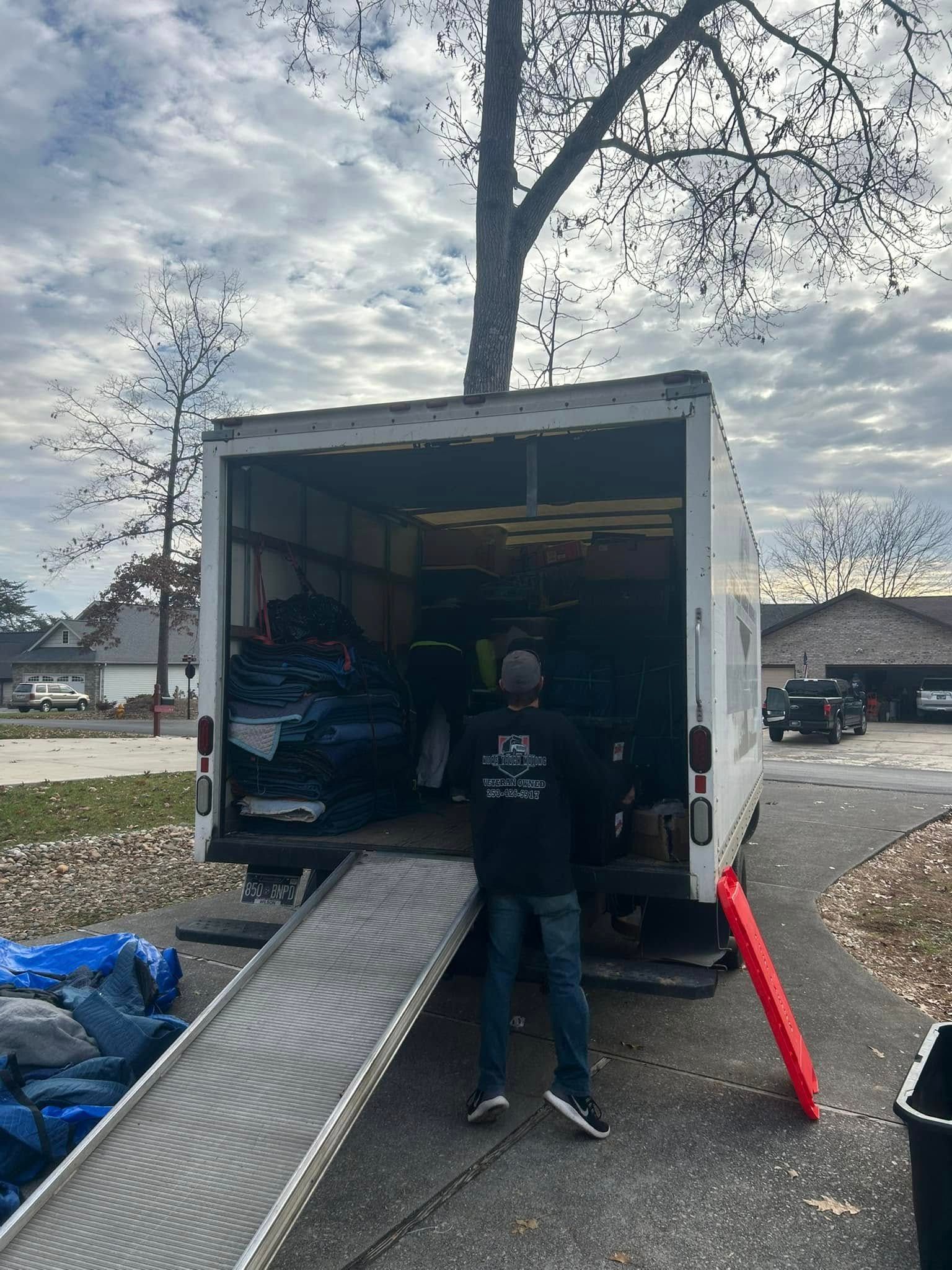 Moving truck open, ramp extended, person stands at back, driveway.