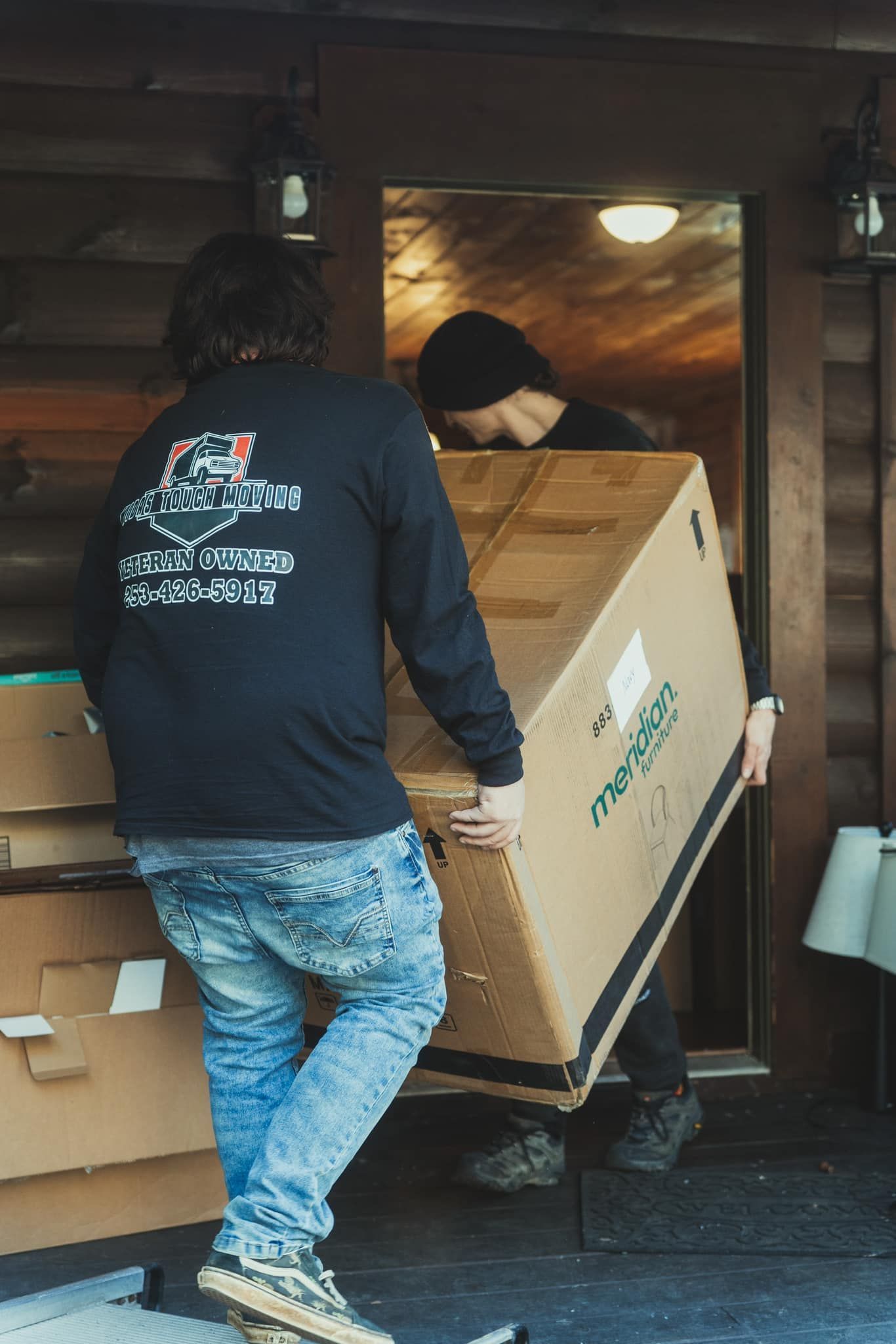 Two people carrying a large cardboard box through a doorway, boxes stacked on the porch.