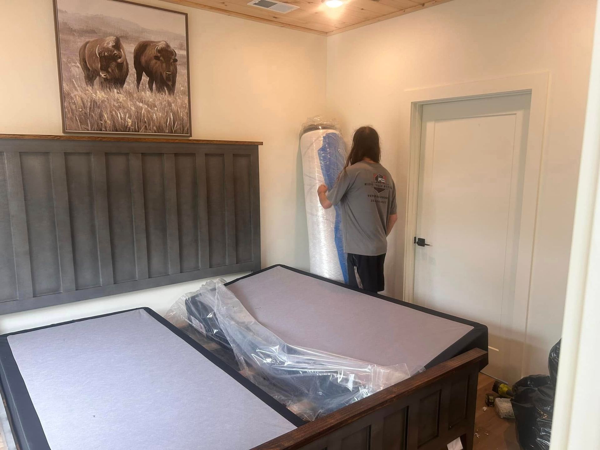 Person holding rolled-up mattress in bedroom with new bed frame and bison art.