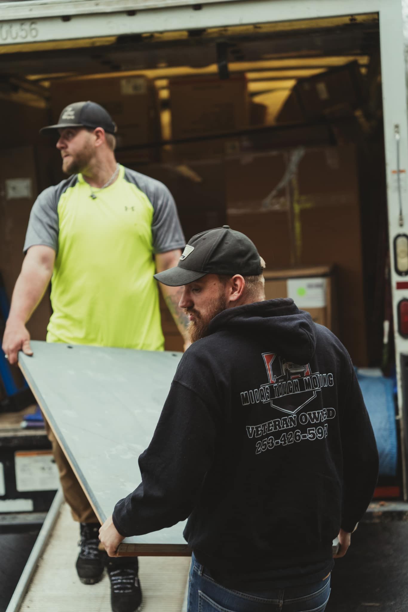 Two men loading a large, gray board into a truck. One wears a black hoodie, the other a yellow shirt.