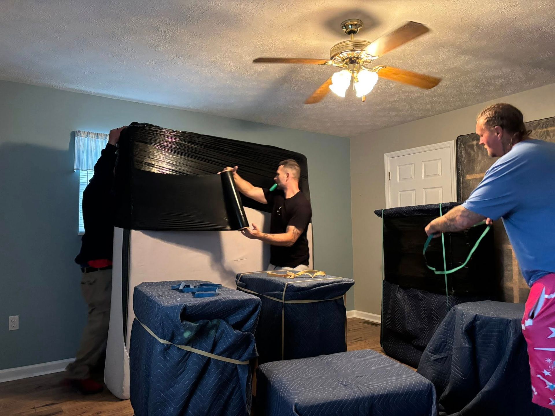 Three people wrapping furniture in a bedroom. One person is securing a mattress. Others tie down wrapped items.
