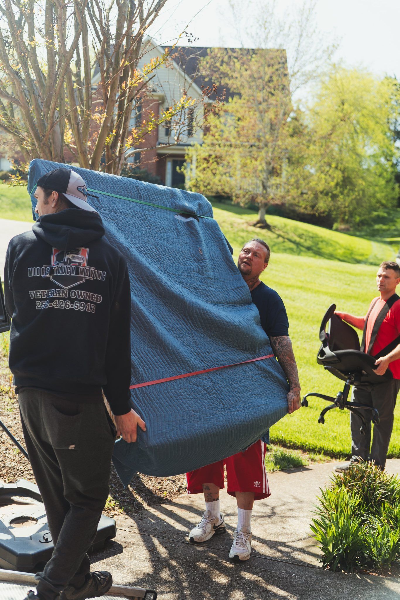 Three people moving furniture outside a house; one carries a large covered mattress, another carries an office chair.