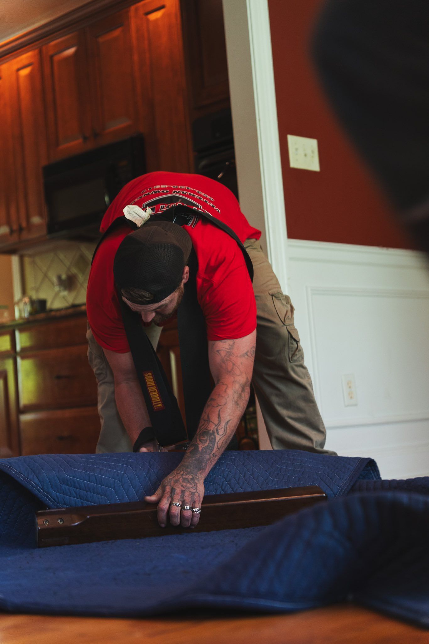 Man in red shirt wrapping a wood object with blue moving blanket in a home.