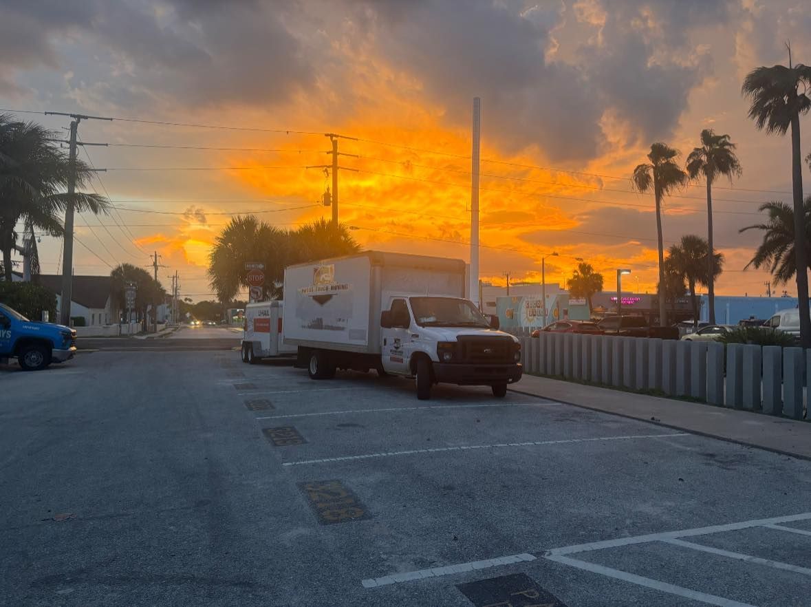 A commercial truck parked on a street with a vibrant orange and yellow sunset. Palm trees and buildings in the background.