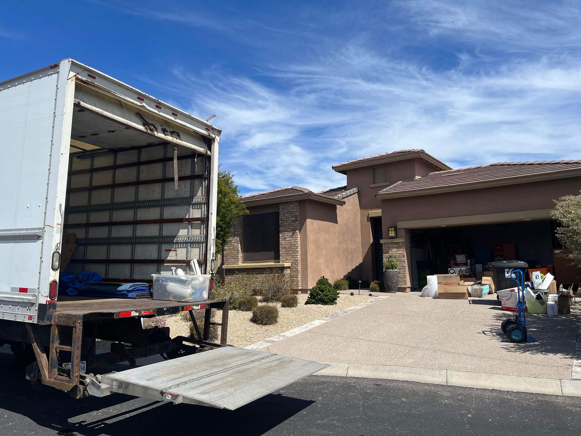 Moving truck parked in front of a house, ramp down. Boxes and items loaded onto the driveway. Blue sky.