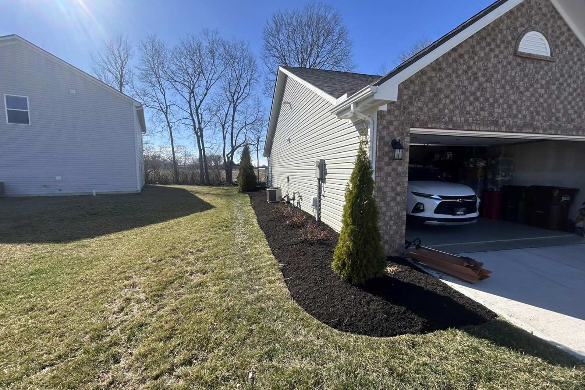 A suburban house with a garage, landscaping, and a car inside. Trees line the side of the house, and the sky is blue.