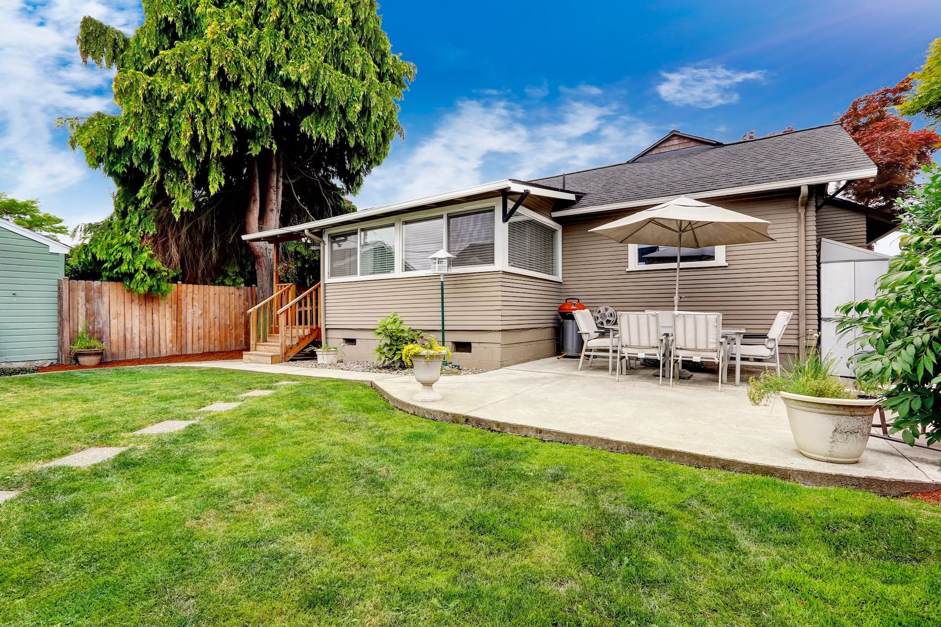 Backyard with a small house, patio with table and chairs, and a green lawn under a blue sky.