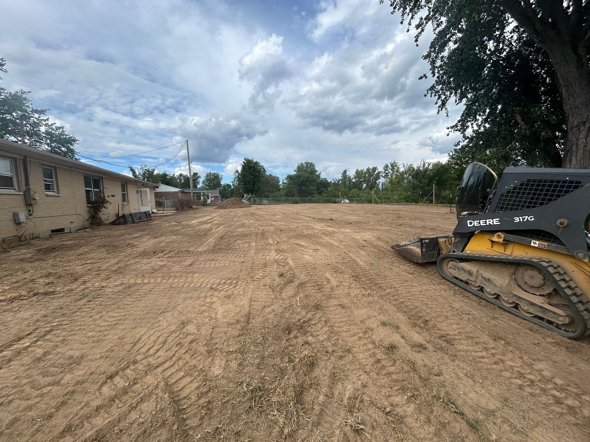 A cleared lot with a skid steer on the right; a low building is on the left. The sky is cloudy.