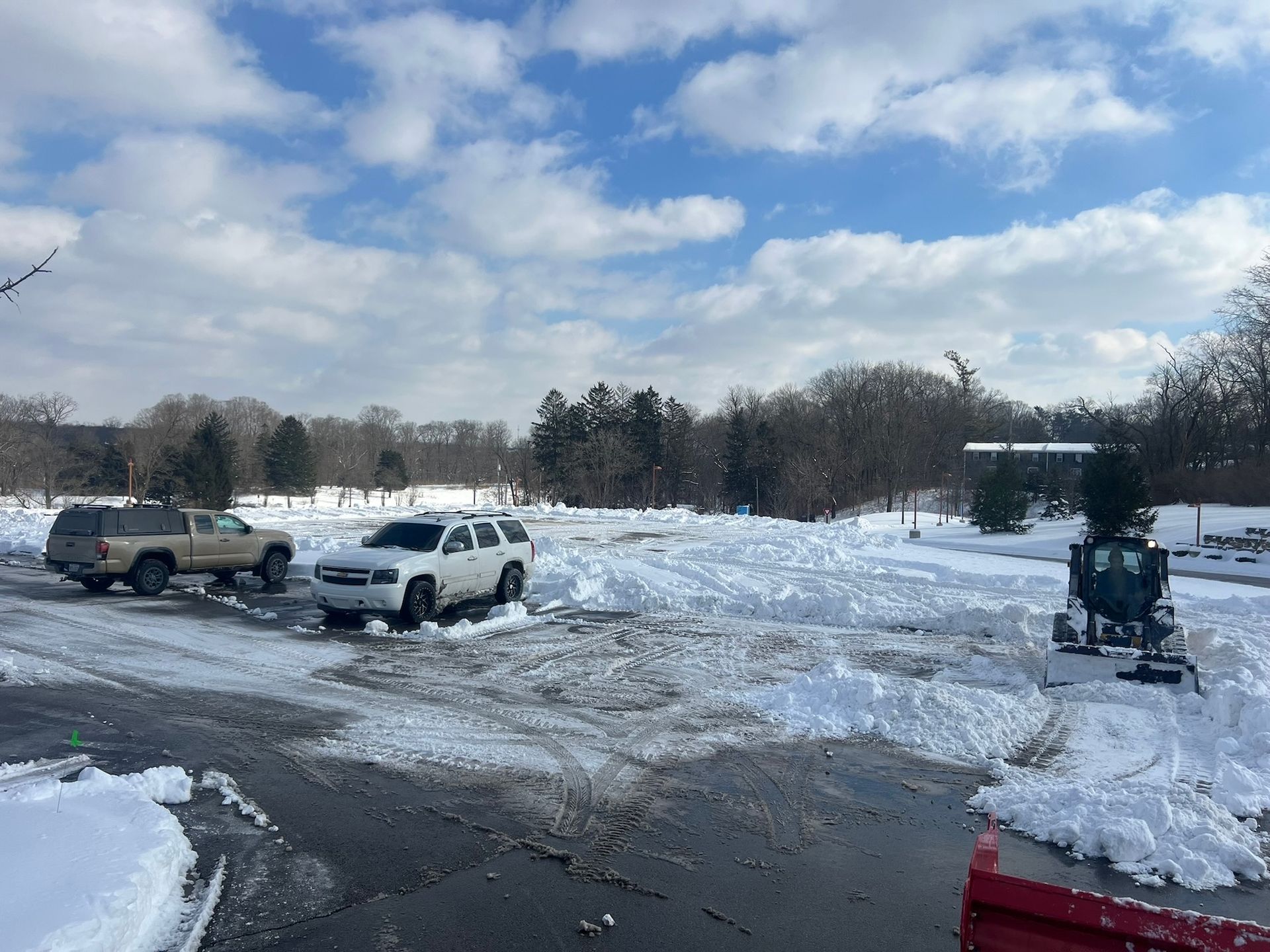 Vehicles in a snow-covered parking lot; a small plow clears snow. Blue sky, bare trees in background.