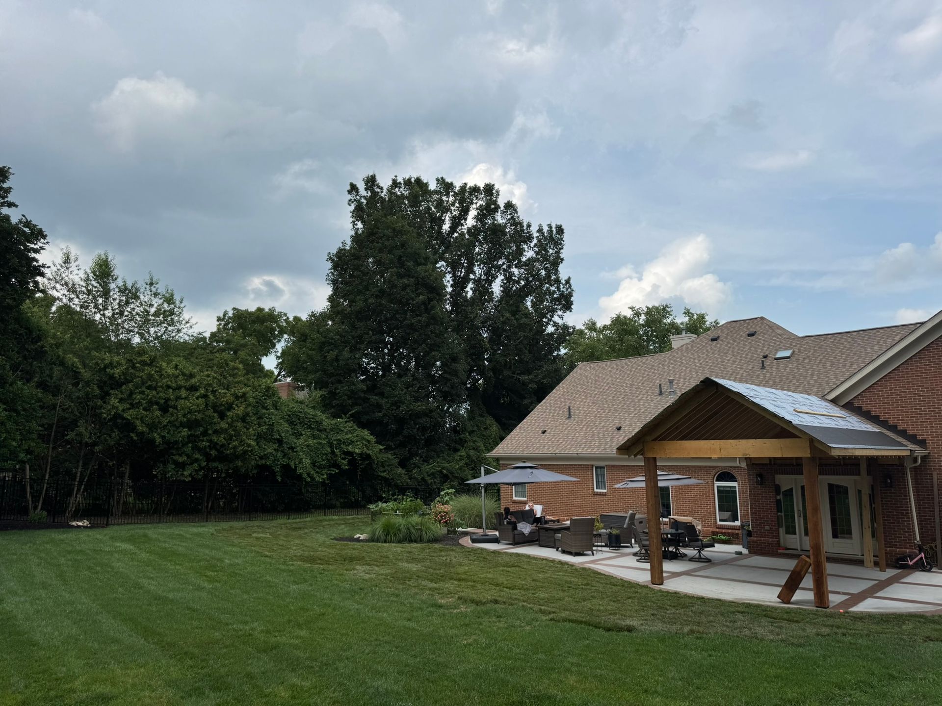 Backyard with brick house, patio, green grass, trees, and cloudy sky.