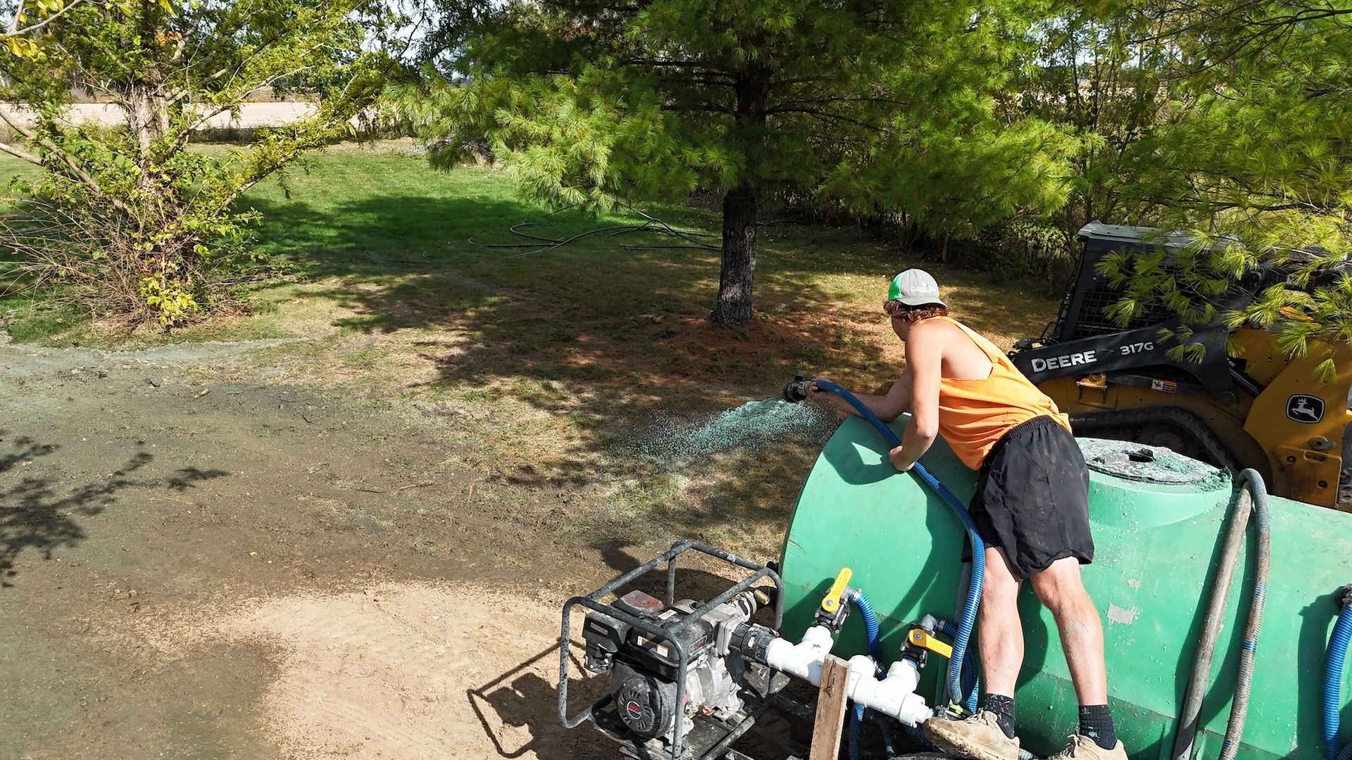 Person spraying water from a green tank on a grassy area near trees.