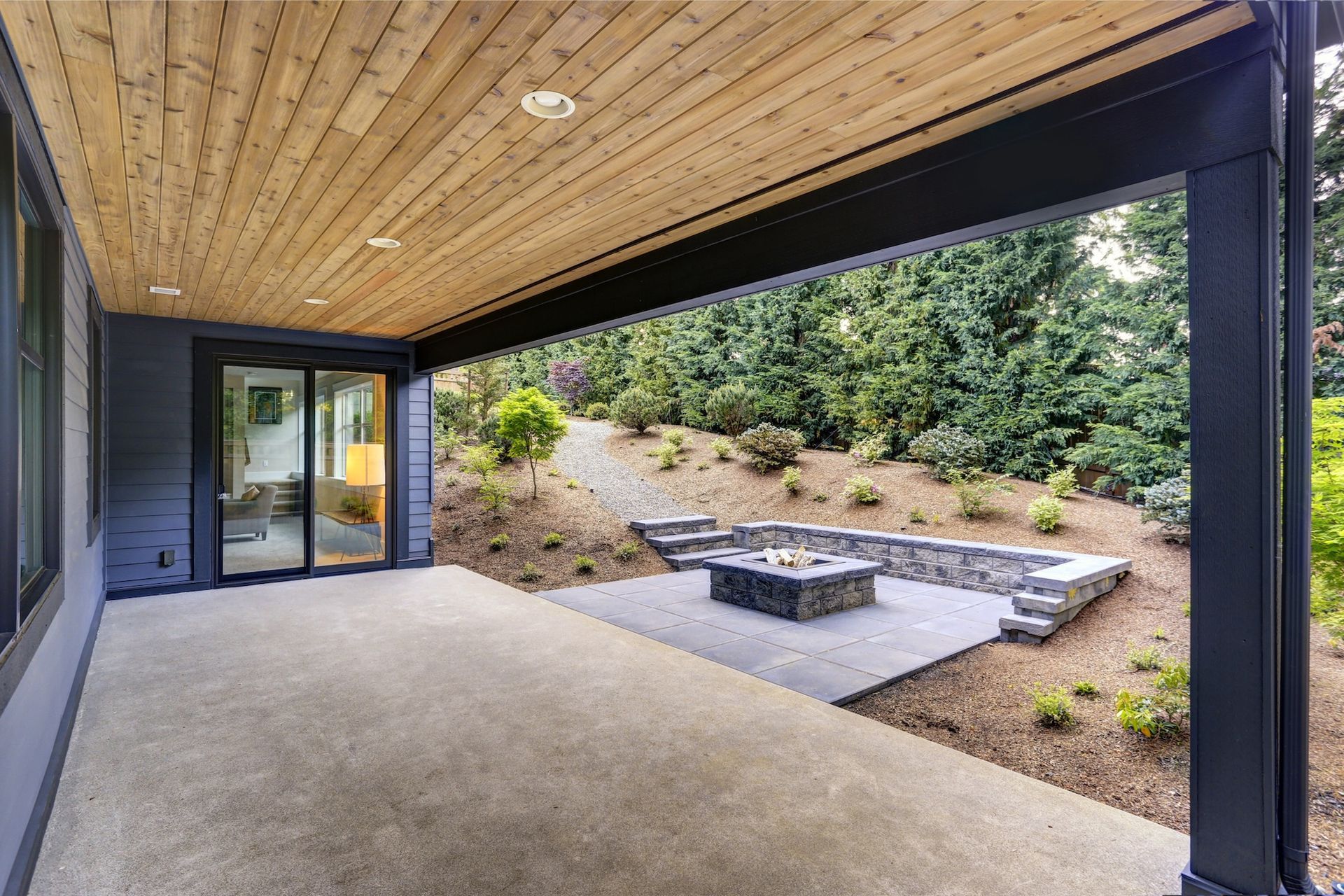 Covered patio with wooden ceiling, overlooking backyard with fire pit and greenery.