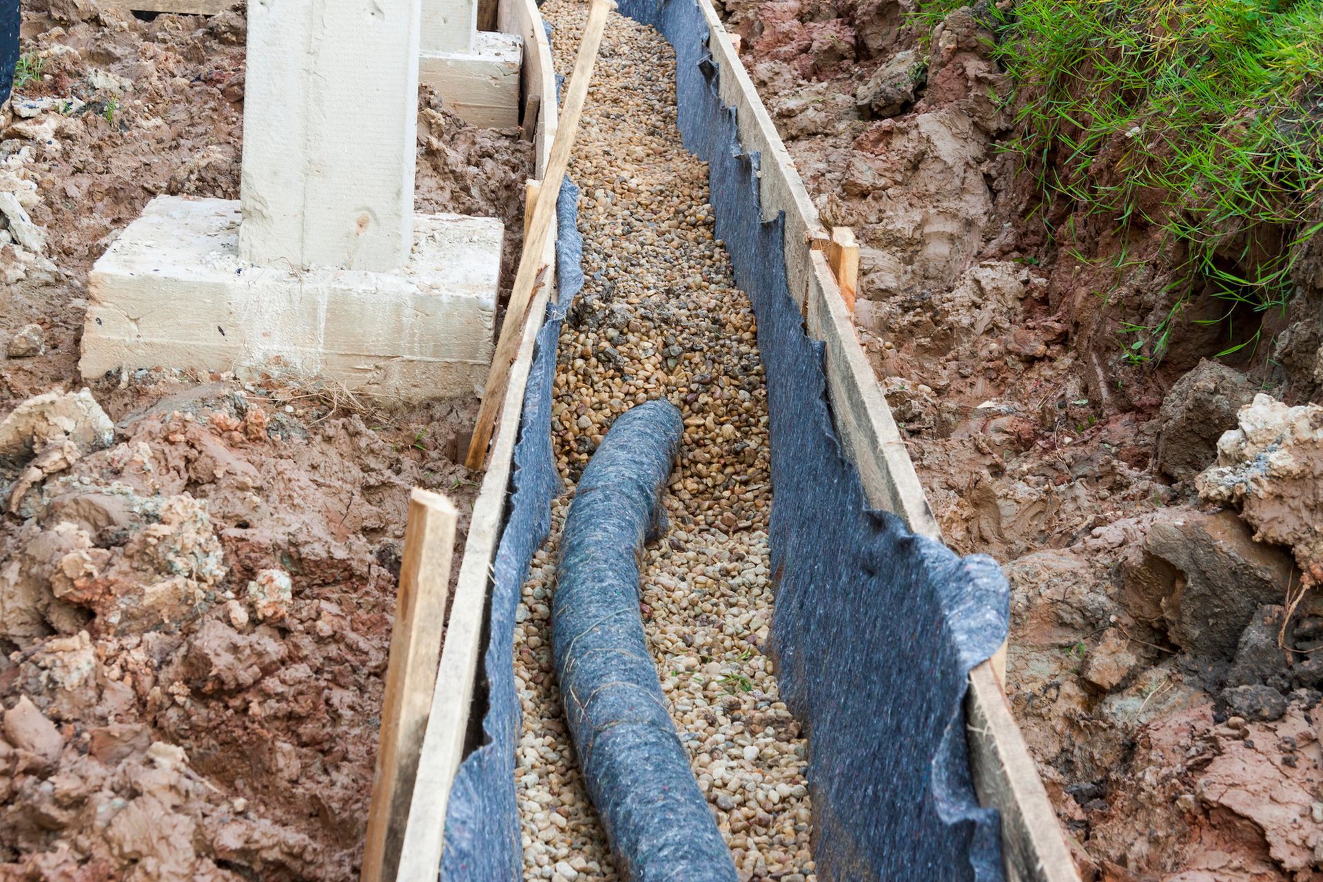 Drainage system installation: trench with gravel, fabric liner, and pipe near a concrete pillar.
