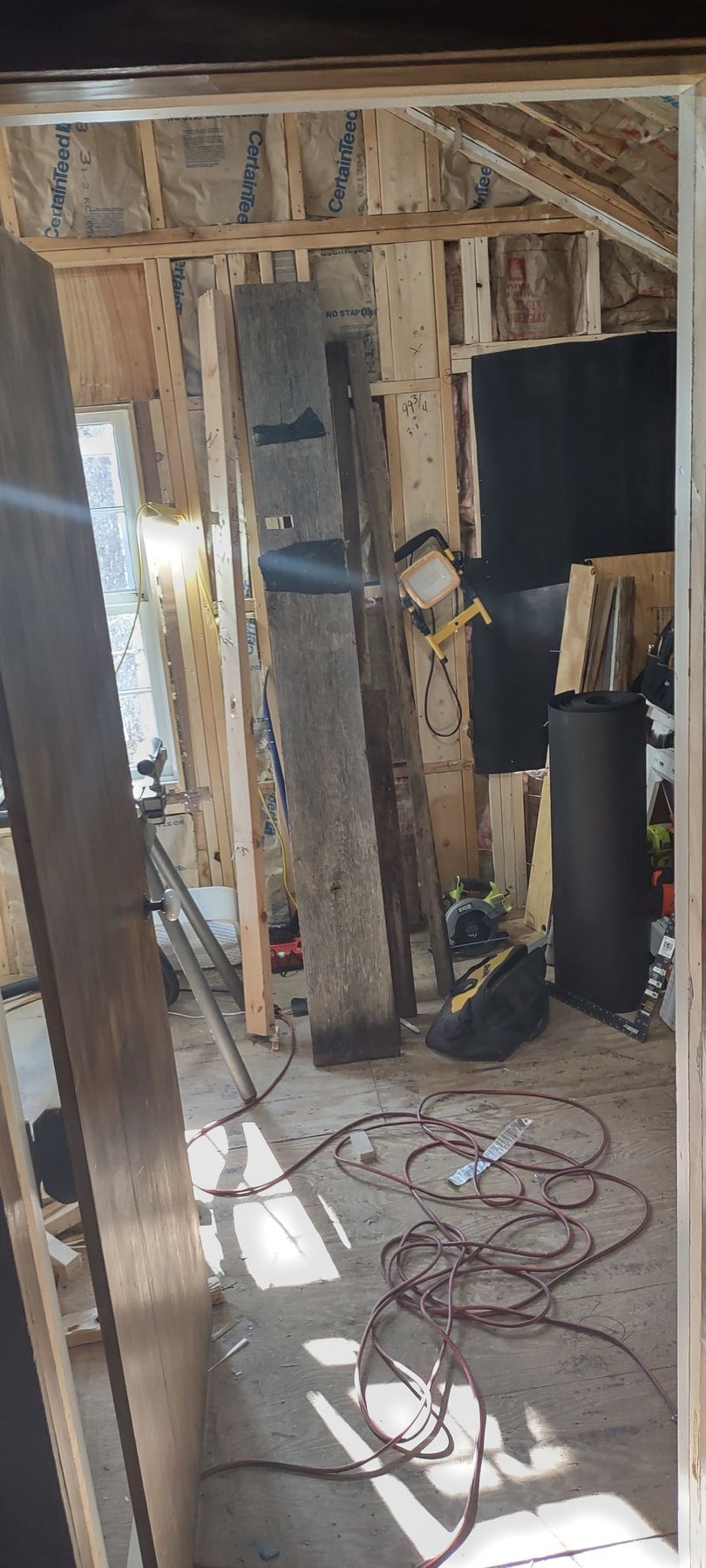 Interior of a room under construction with exposed wooden beams, a tall pillar, tools, and electrical cords on the floor.