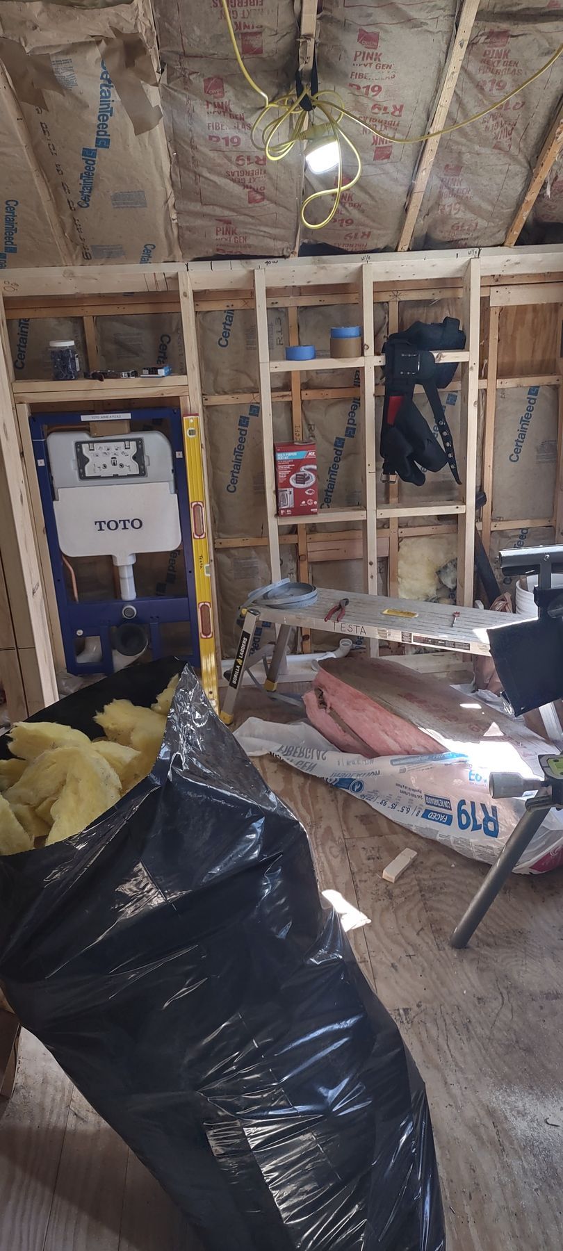 Interior construction site with exposed framing, insulation, a black trash bag, and a toilet fixture.
