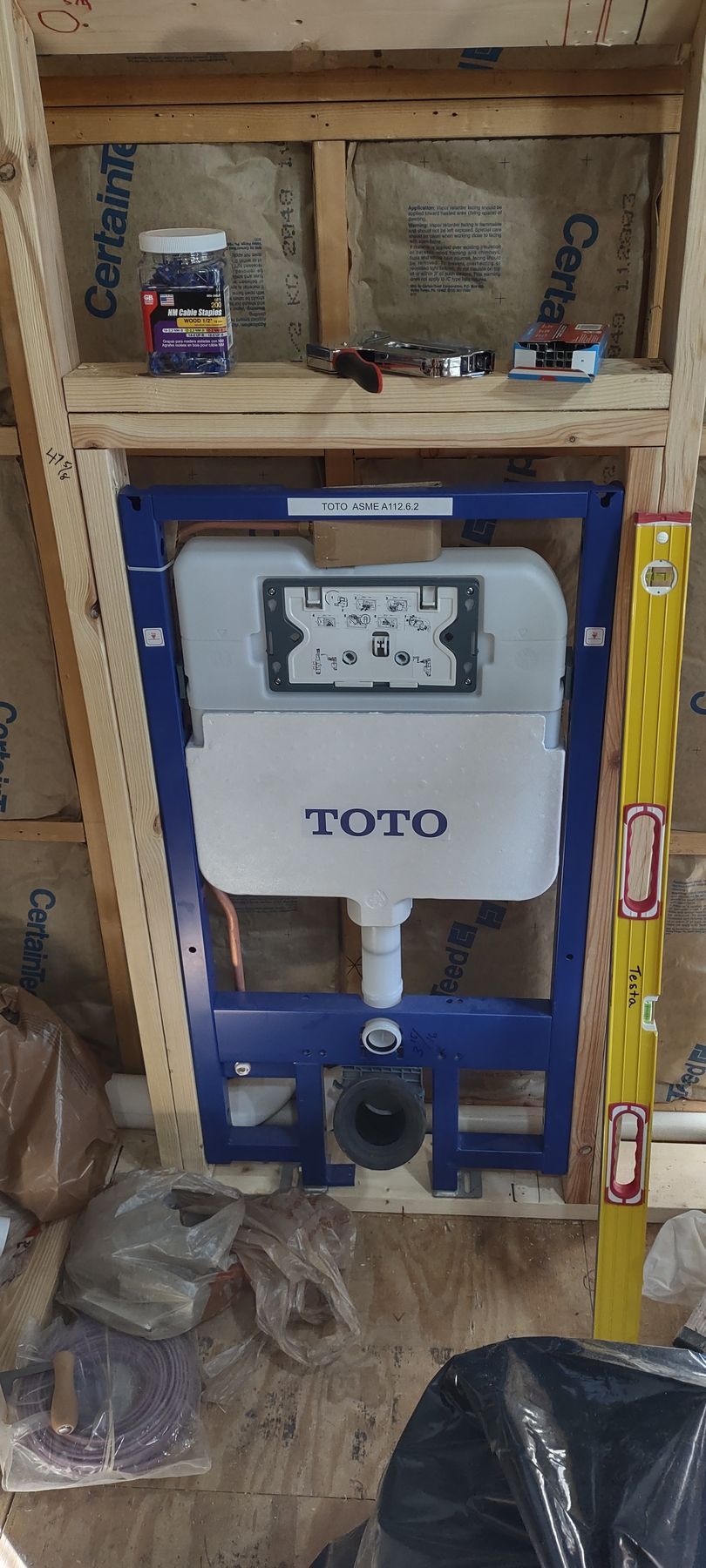 A blue TOTO toilet tank installed in a wooden frame with a level. Tools and materials are on a shelf above.