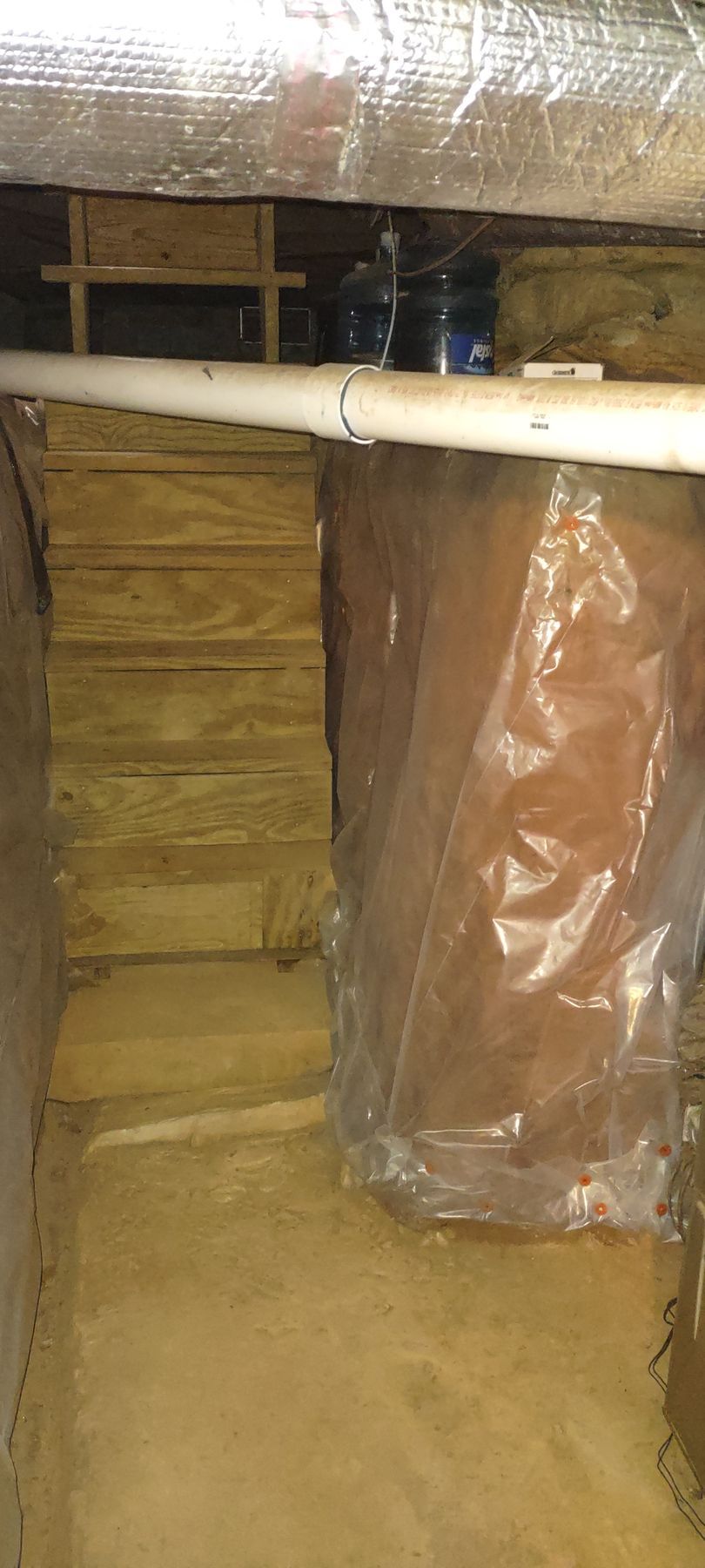 Wooden stairs in a basement, with a plastic barrier covering a support beam and a white pipe overhead.