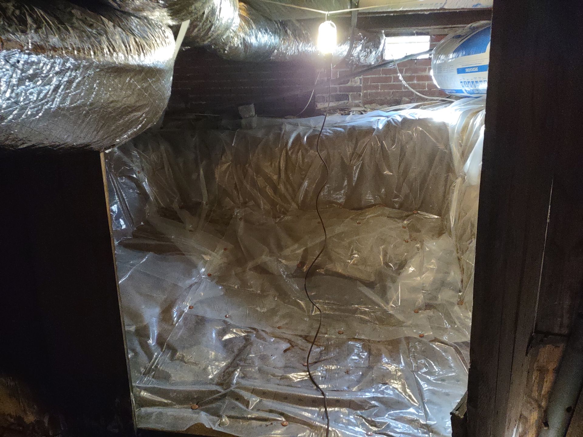 Crawlspace with plastic sheeting covering a wall, insulation above. Sunlight streams in.