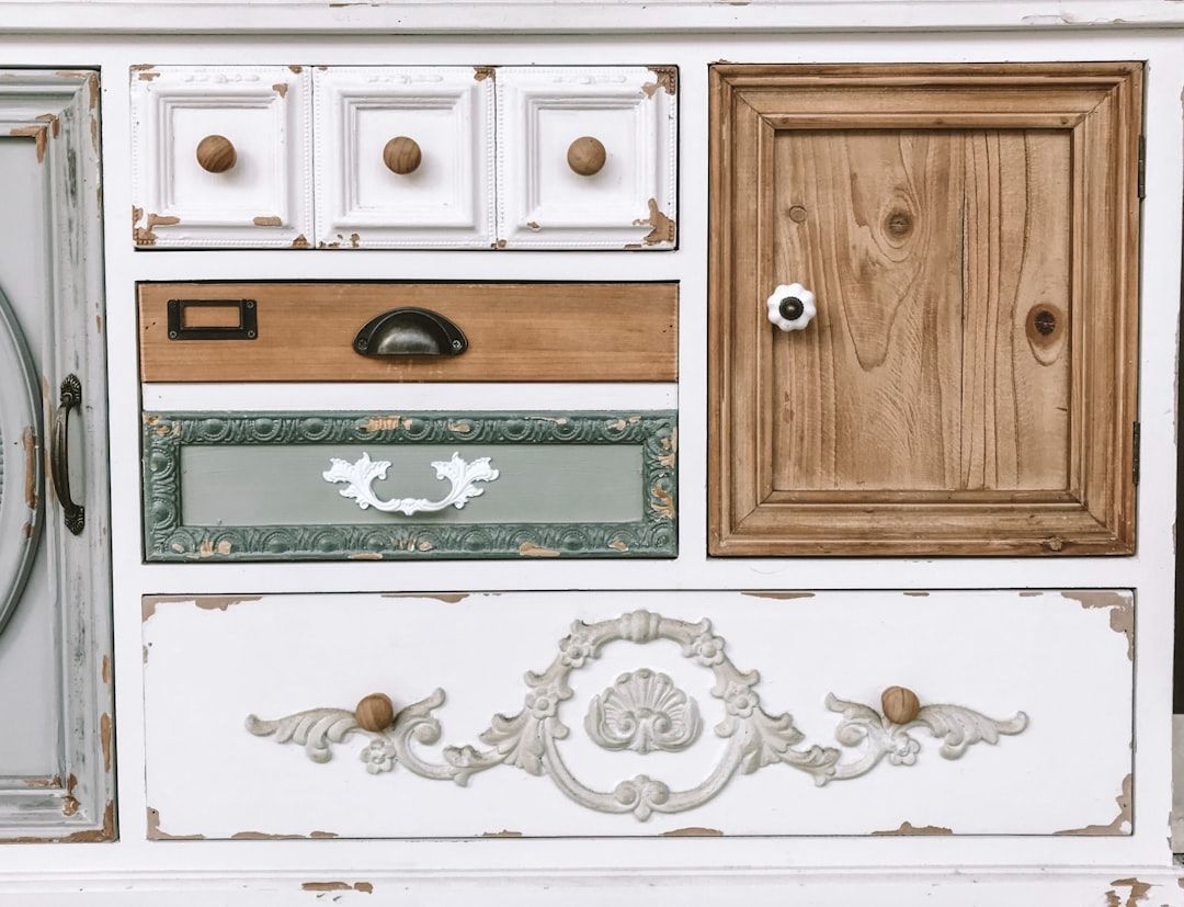 Close-up of a white, distressed cabinet with various drawers and a wooden door in contrasting colors. Decorative carvings add detail.