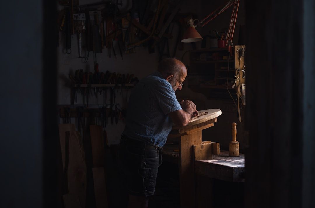 Elderly man working at a workbench in a dimly lit workshop. He is carving wood; tools hang on the wall behind him.