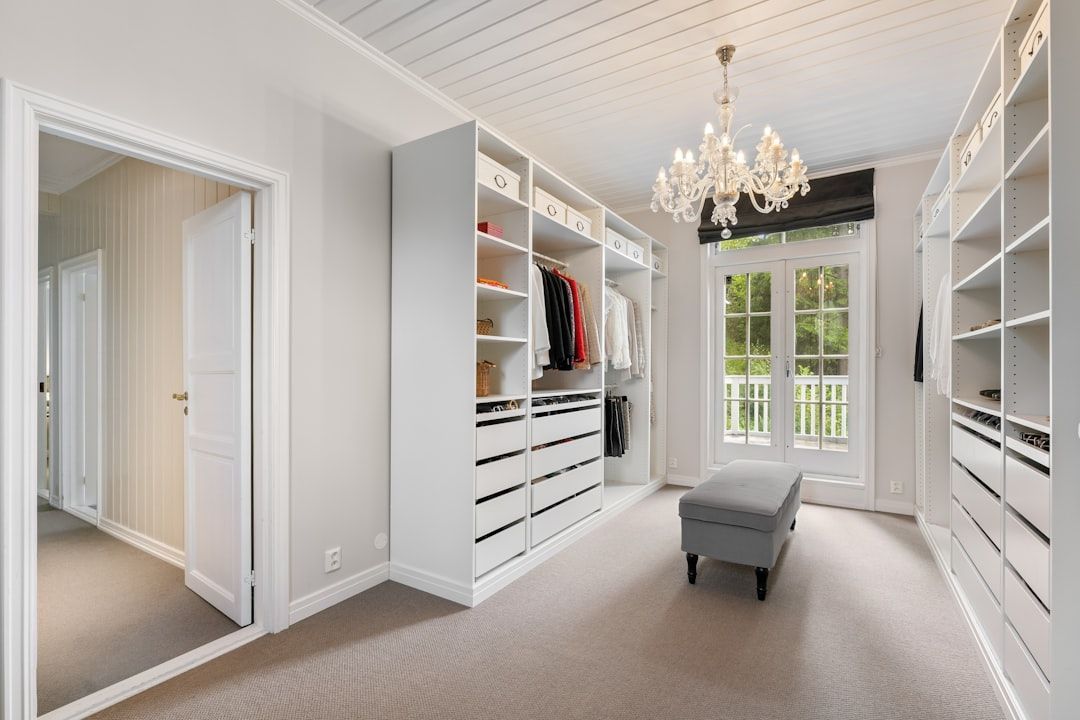 Walk-in closet with white built-in shelving, drawers, and a chandelier. Features a gray ottoman and a doorway leading outside.