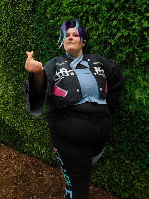 A queer, nonbinary, and plus-size person in a Sombra cosplay. They are posed upright and are making a small heart symbol with their fingers. 