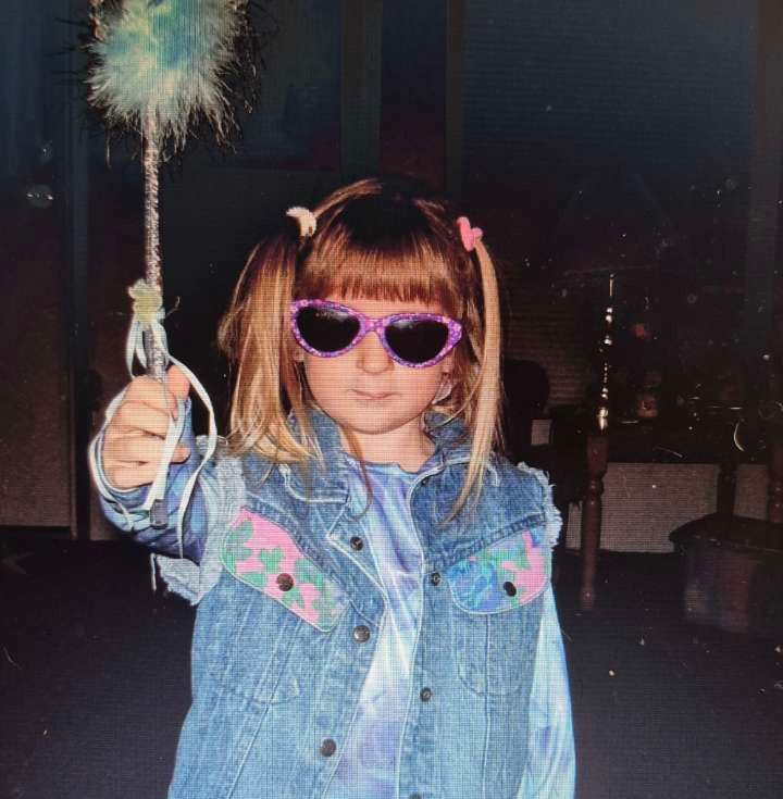 A young, autistic child with cleft lip & palate. The child is wearing large, purple, cat-eye glasses and a light blue jean jacket. They are holding a light blue fuzzy wand towards the camera with a straight face.